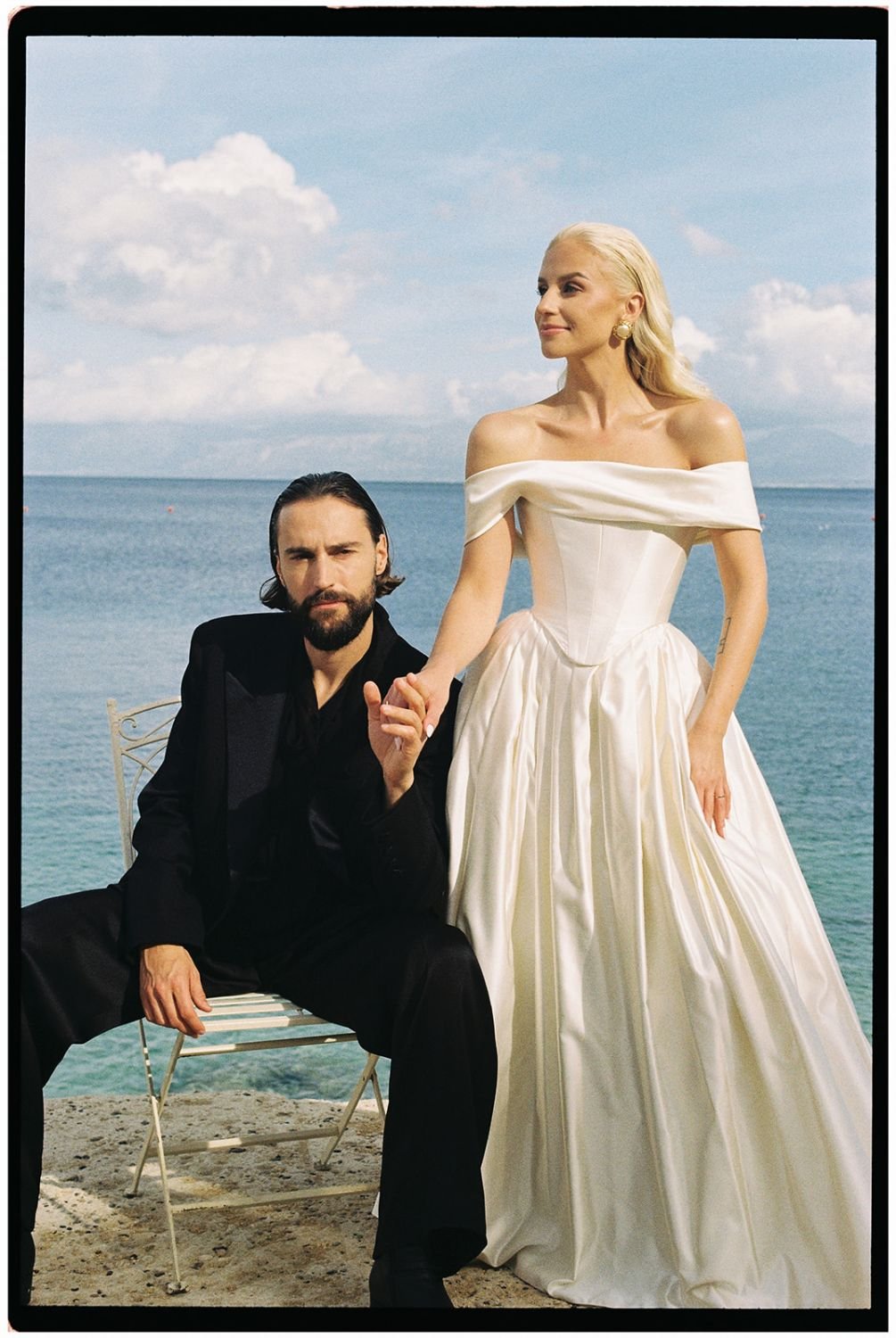 A man in a black suit sitting on a white chair by the ocean, holding hands with a woman in a white off-the-shoulder gown. The background shows a blue sky with clouds and the sea.