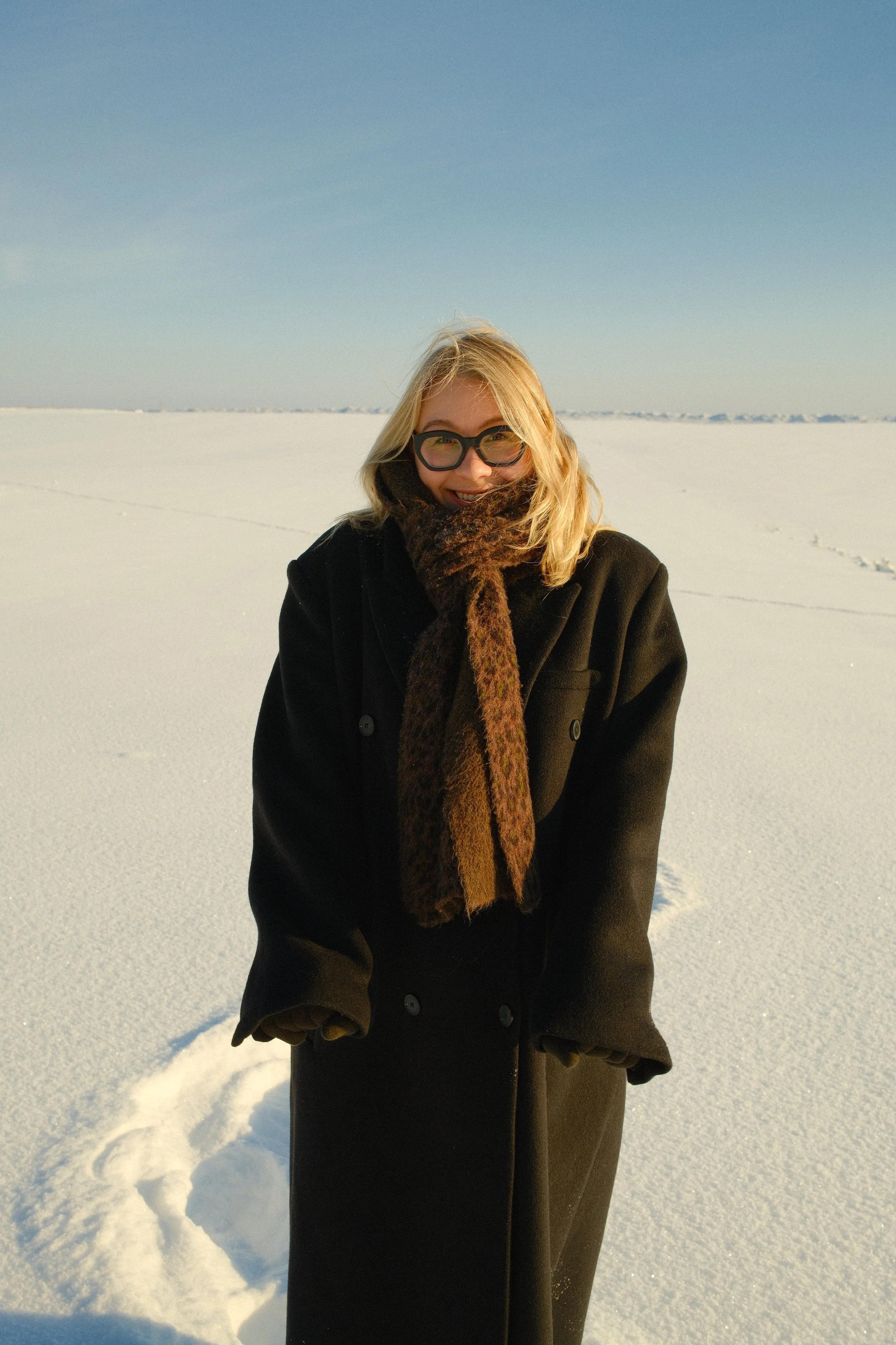 Woman in black coat and animal print scarf standing in snow-covered landscape with clear blue sky.