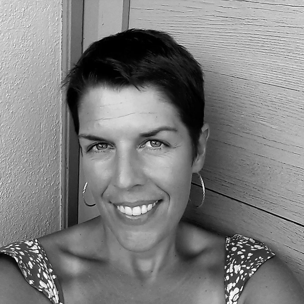 Schwarz-weiß Porträt einer Frau mit kurzem dunklem Haar, großen Ohrringen, lächelnd, in einem sommerlichen Outfit, vor einer Holz- und Putzwand