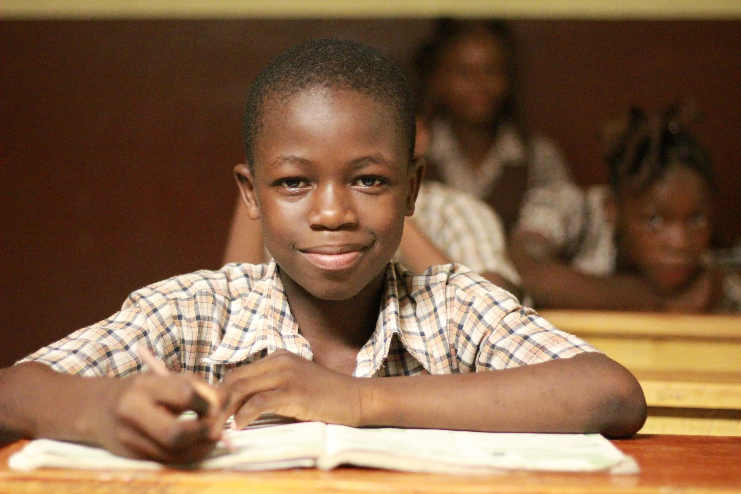 Young Haitian boy in a classroom smiling as he writes in his notebook while other students are working in the background