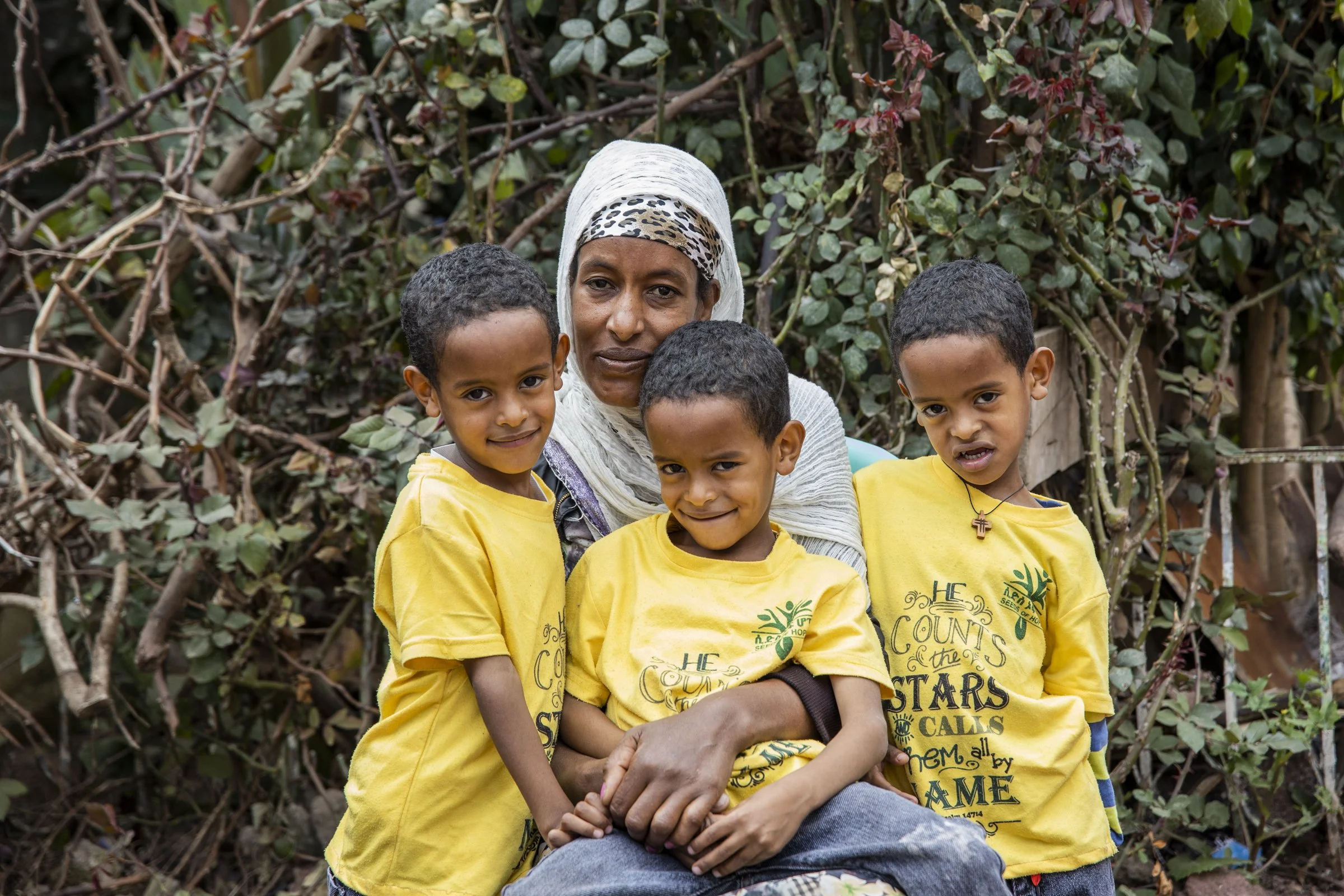 An Ethiopian mom smiles with her three little triplet boys, she holds one on her lap as the other two stand on both sides of her in front of greenery