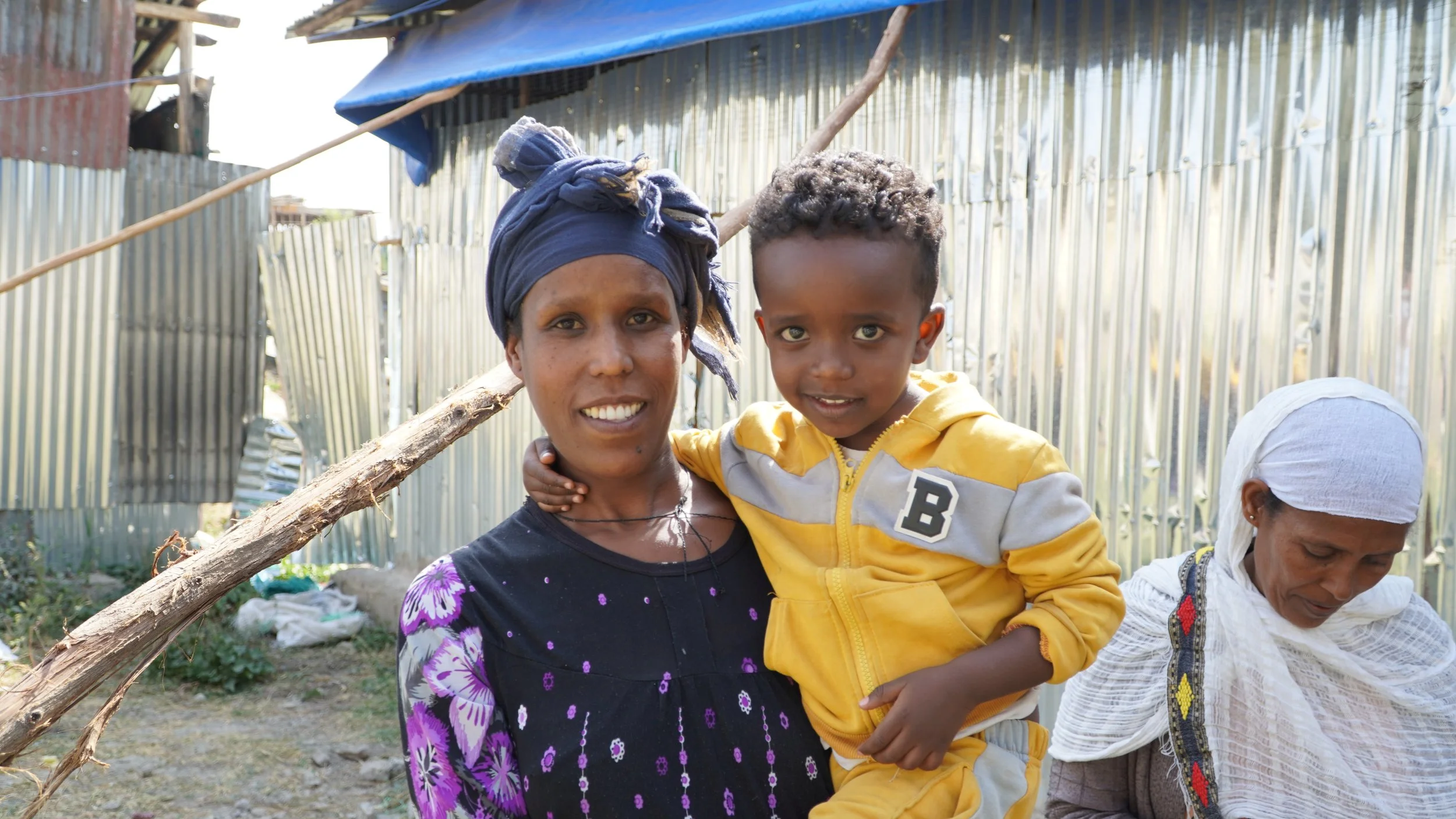 Ethiopian mother holding her little son as they both smile outside in front of a tin building