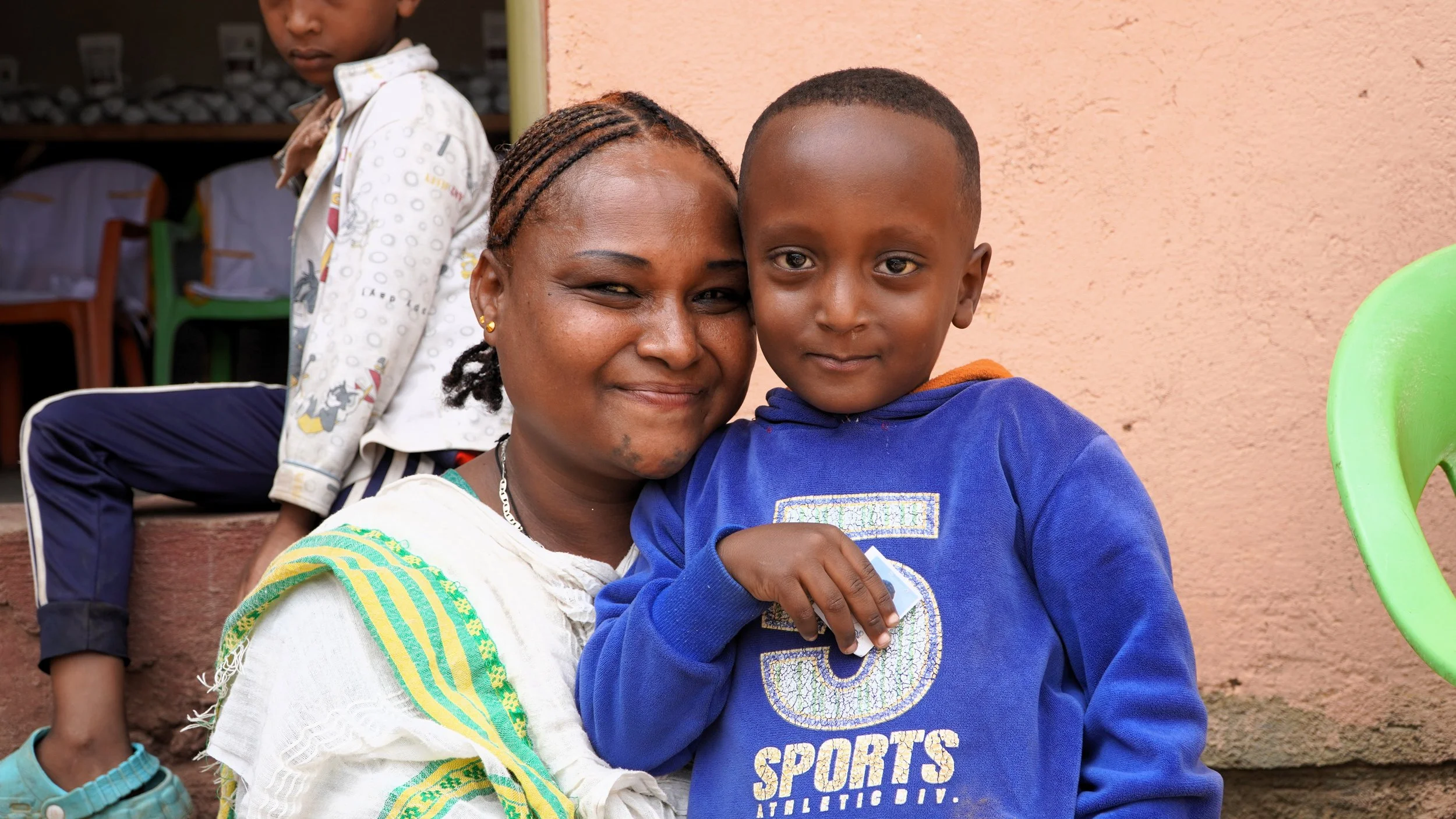 An Ethiopian mom holds her little boy close and they smile outside in front of a cement building