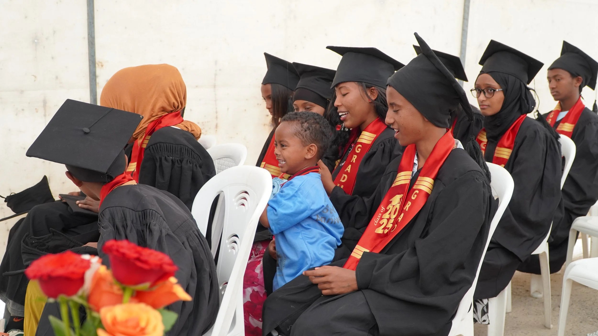 A group of children are seated and wearing caps and gowns, a little boy sits on a girls lap and laughs with them