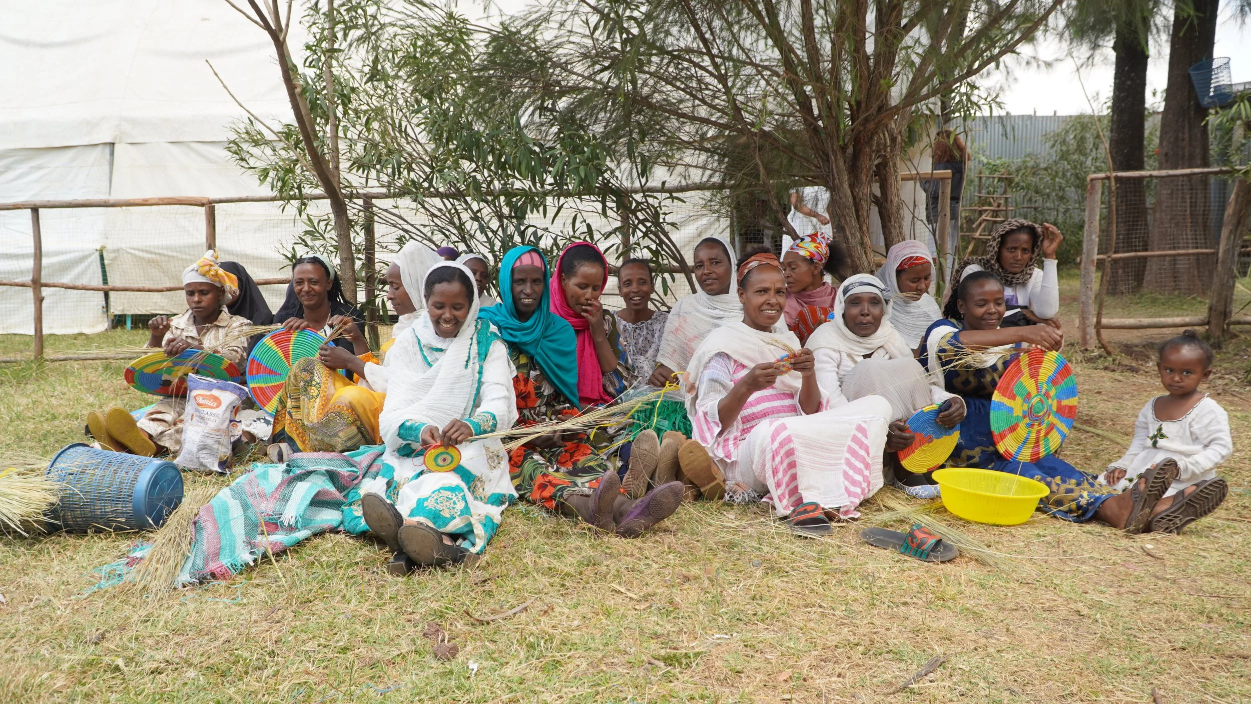 Large group of mothers sitting together outside, smiling as they weave baskets