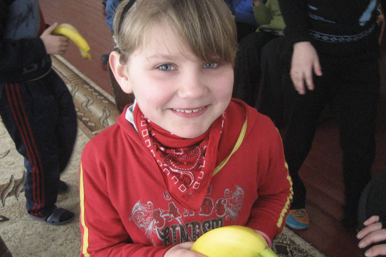 A young Ukrainian girl smiles and holds a banana in a house with other children behind her