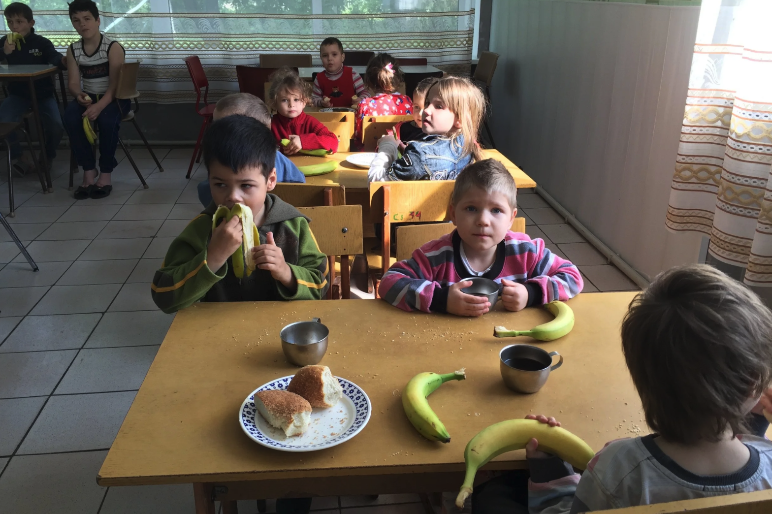 Many Ukrainian children sit at tables eating bananas and bread and drinking water