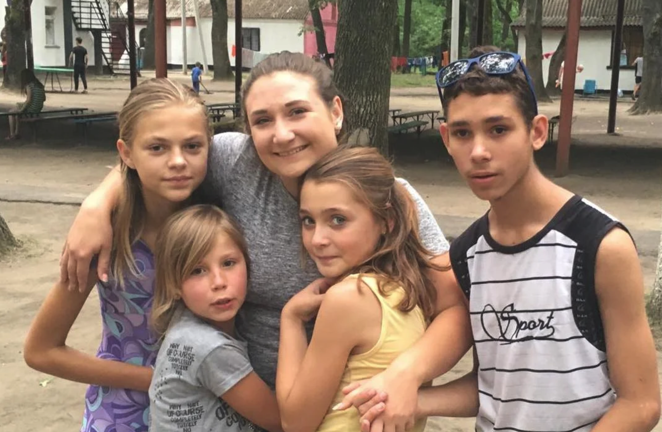 A Ukrainian family with a mom, three daughters, and a son smile and have their arms around each other in a park