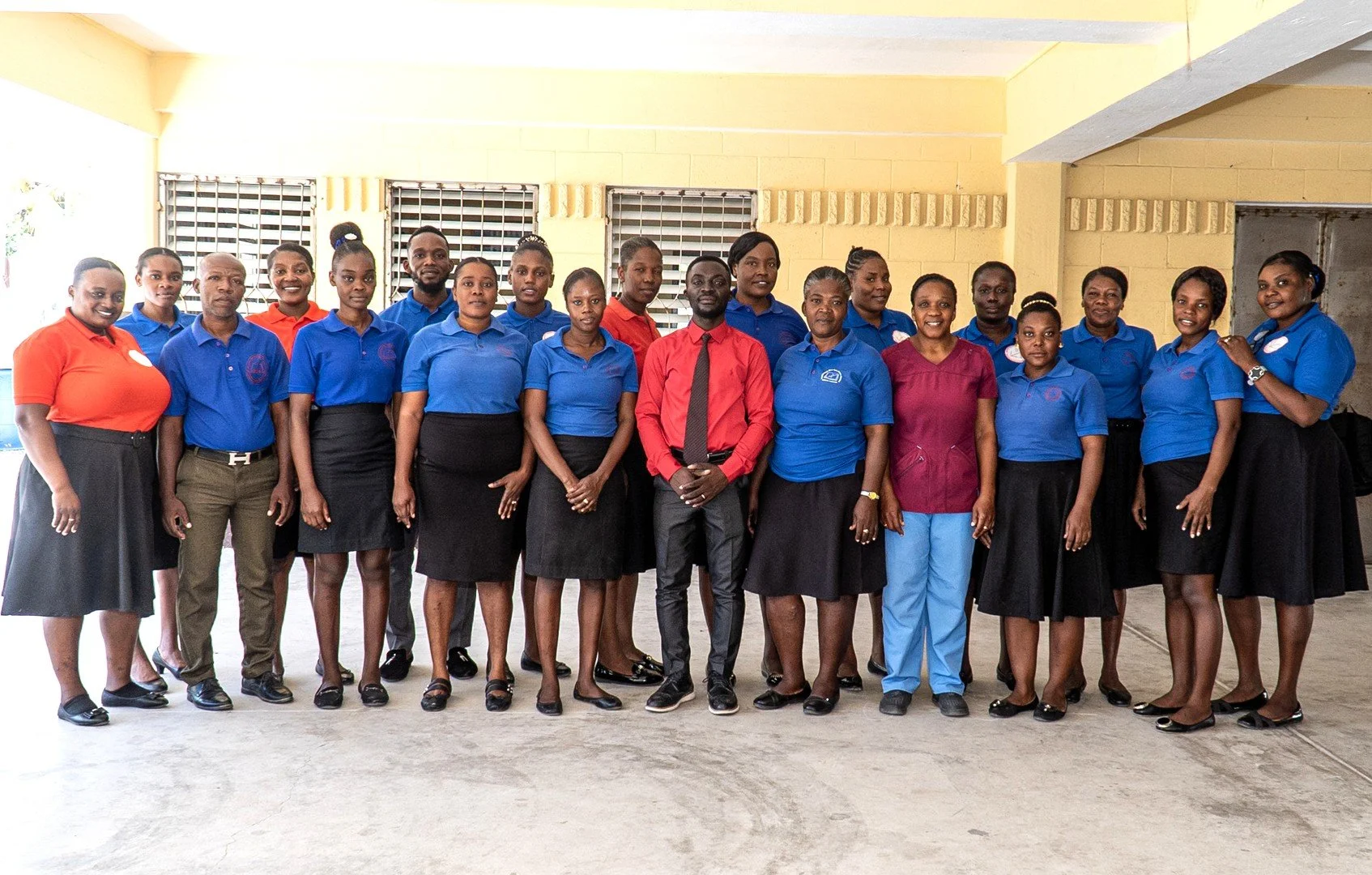 A group of twenty Haitian men and women smiling and standing together in front of a yellow, cement building