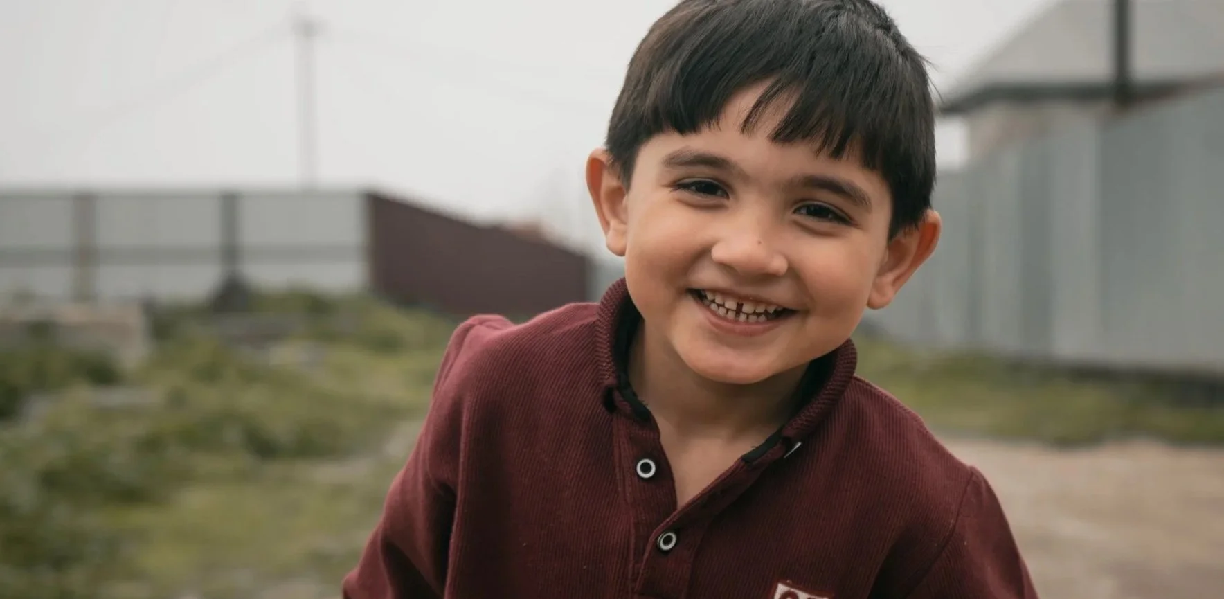 Little Ukrainian boy with dark hair and dark eyes smiling outside on a cloudy day with tin buildings in the background