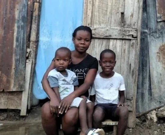A young Haitian mother smiles with her two little boys, one of them sits on her lap and the other sits beside her in front of their home
