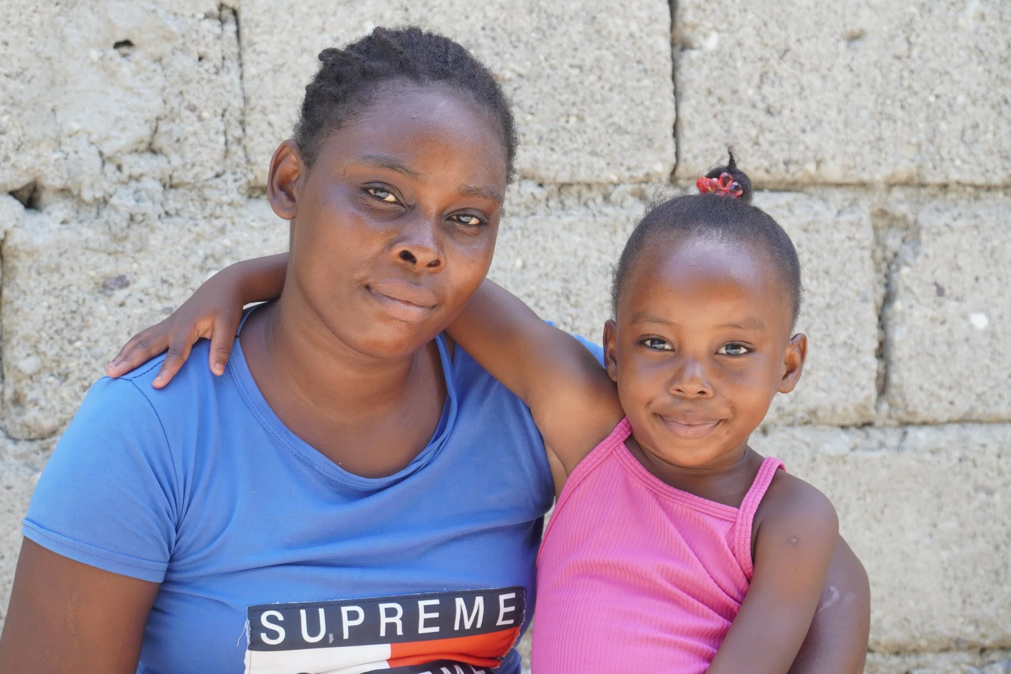 A young Haitian mother smiles with her little girl who has her arm around her in front of a cement building