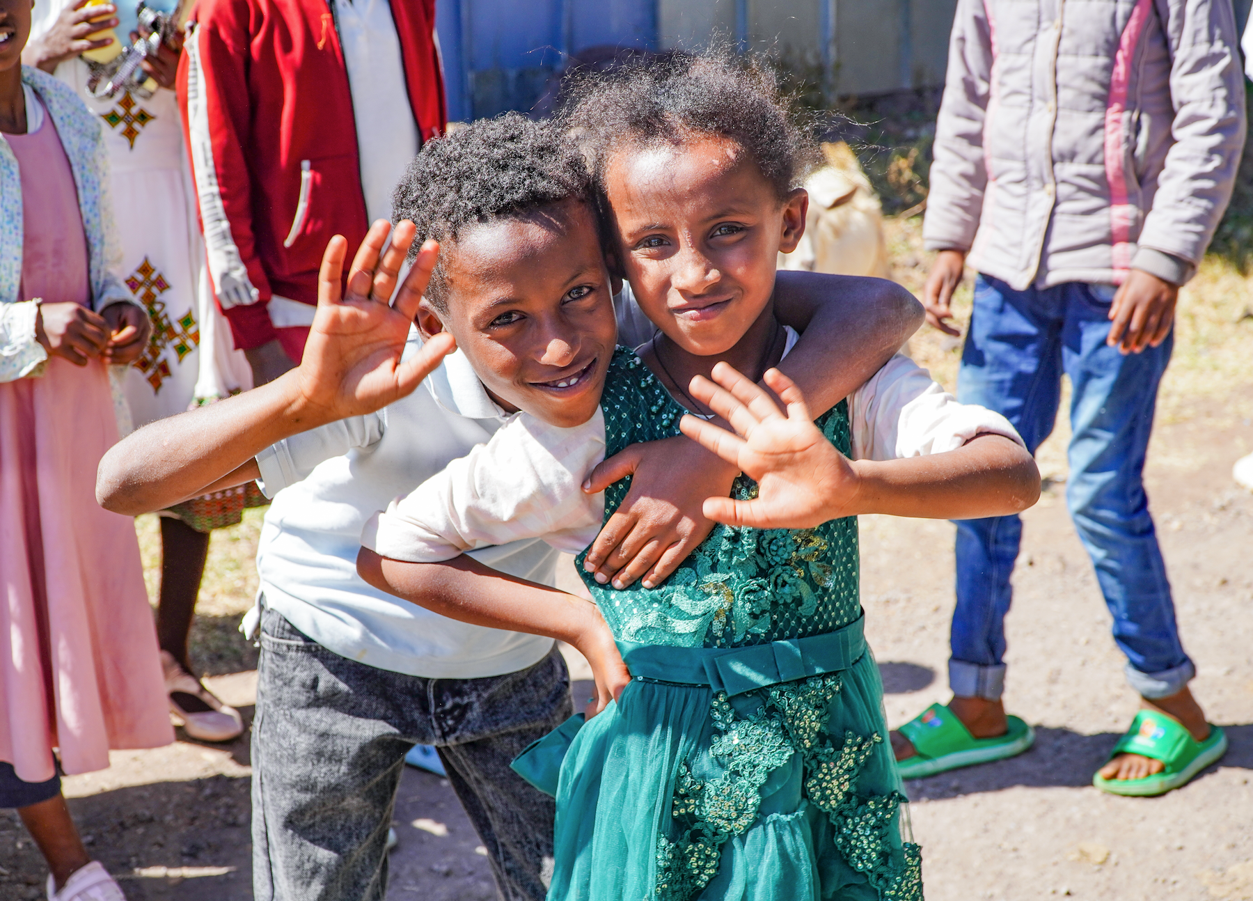A young Ethiopian brother and sister smile with their arms around each other outside with other children behind them