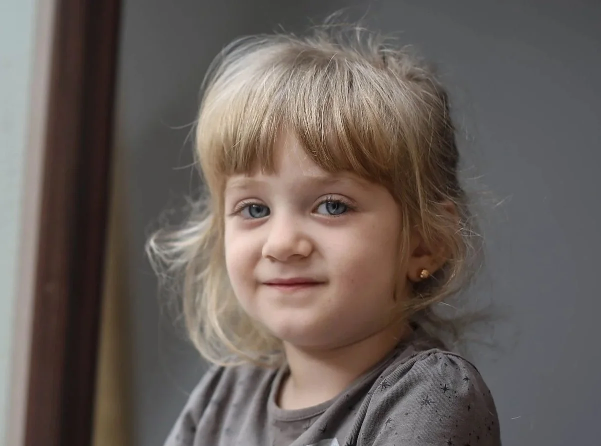 A little Ukrainian girl with blonde hair in a pony tail and blue eyes smiles softly inside her home