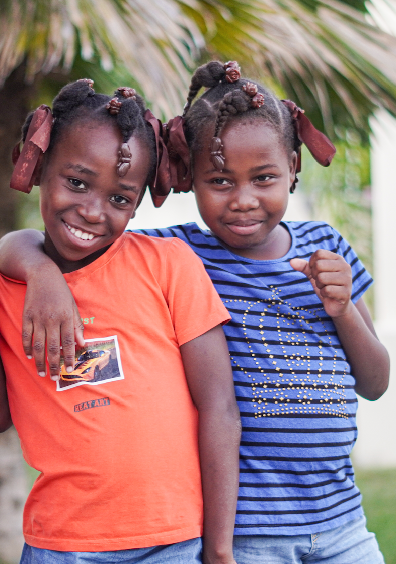 Two young Haitian girls smiling with their arms around each other and greenery in the background