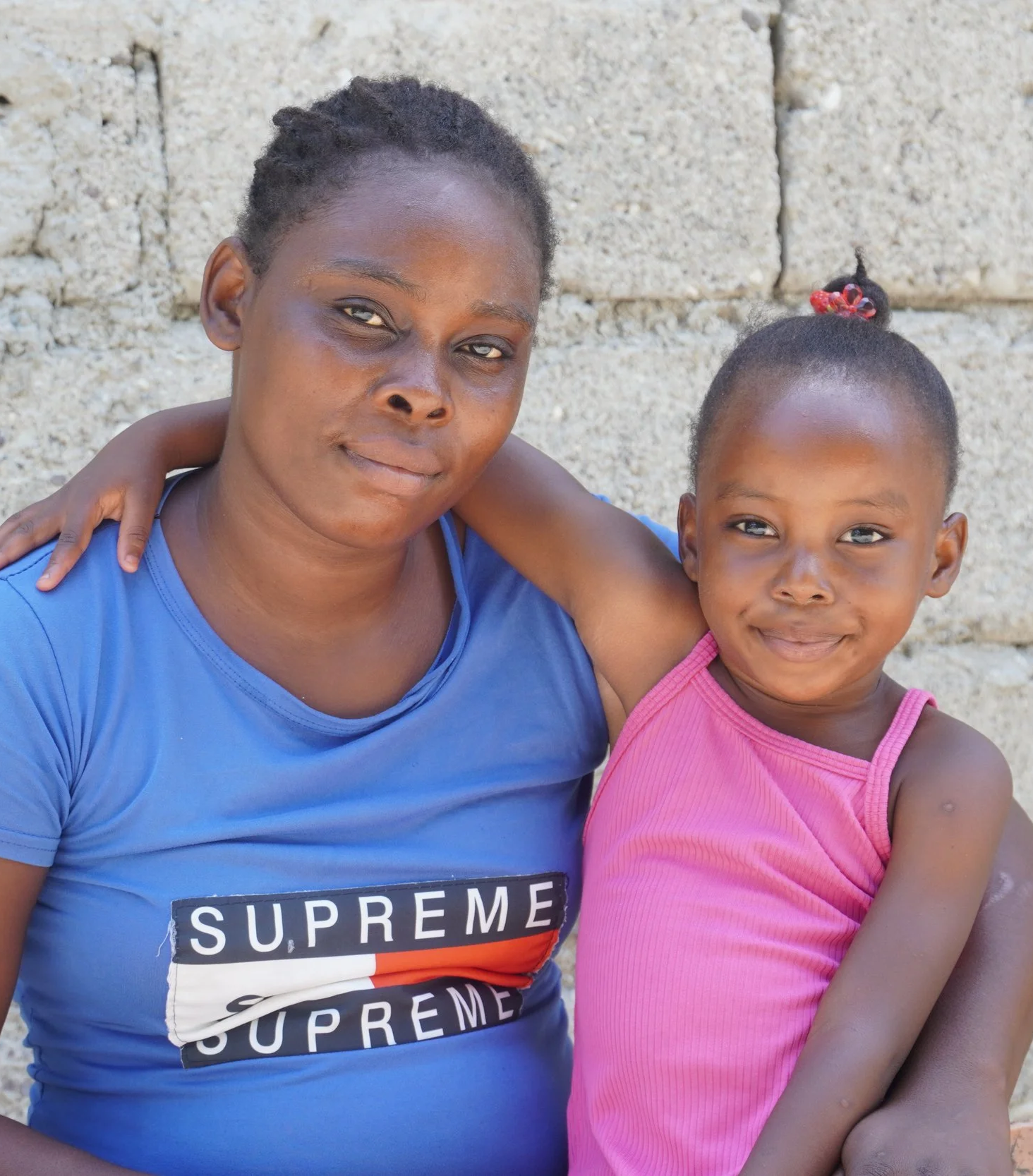 A young Haitian mother smiles with her little girl who has her arm around her in front of a cement building
