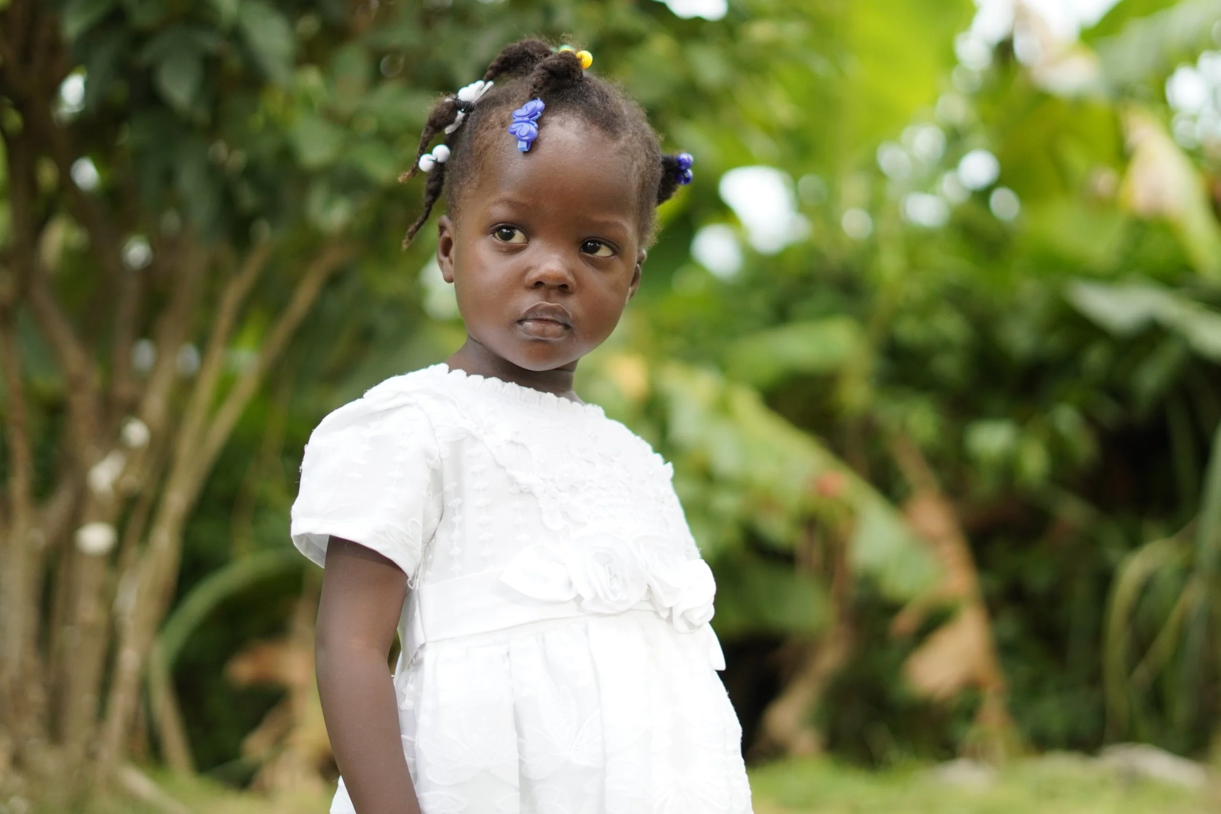 Little Haitian girl in a white dress solemnly looking up with greenery in the background