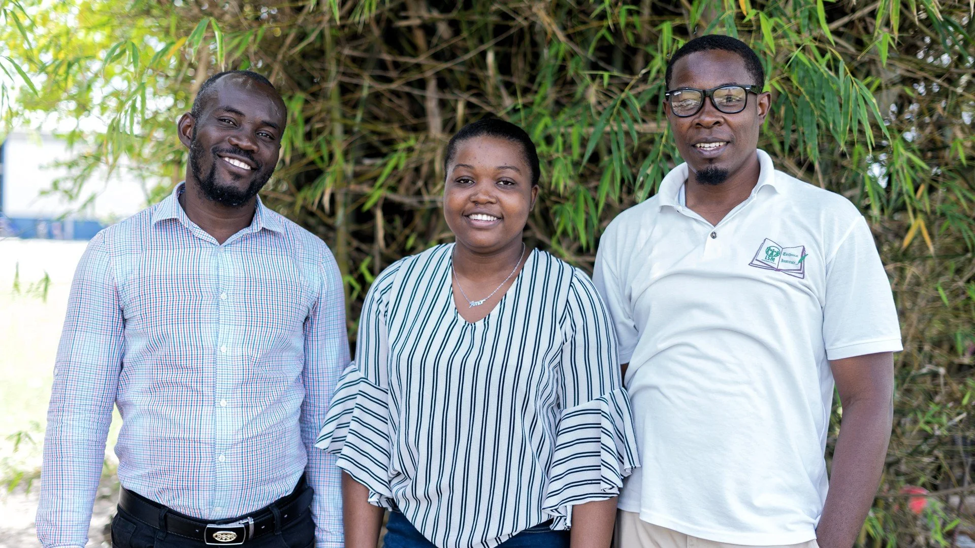 Two men and one woman Haiti staff members standing together smiling in front of greenery