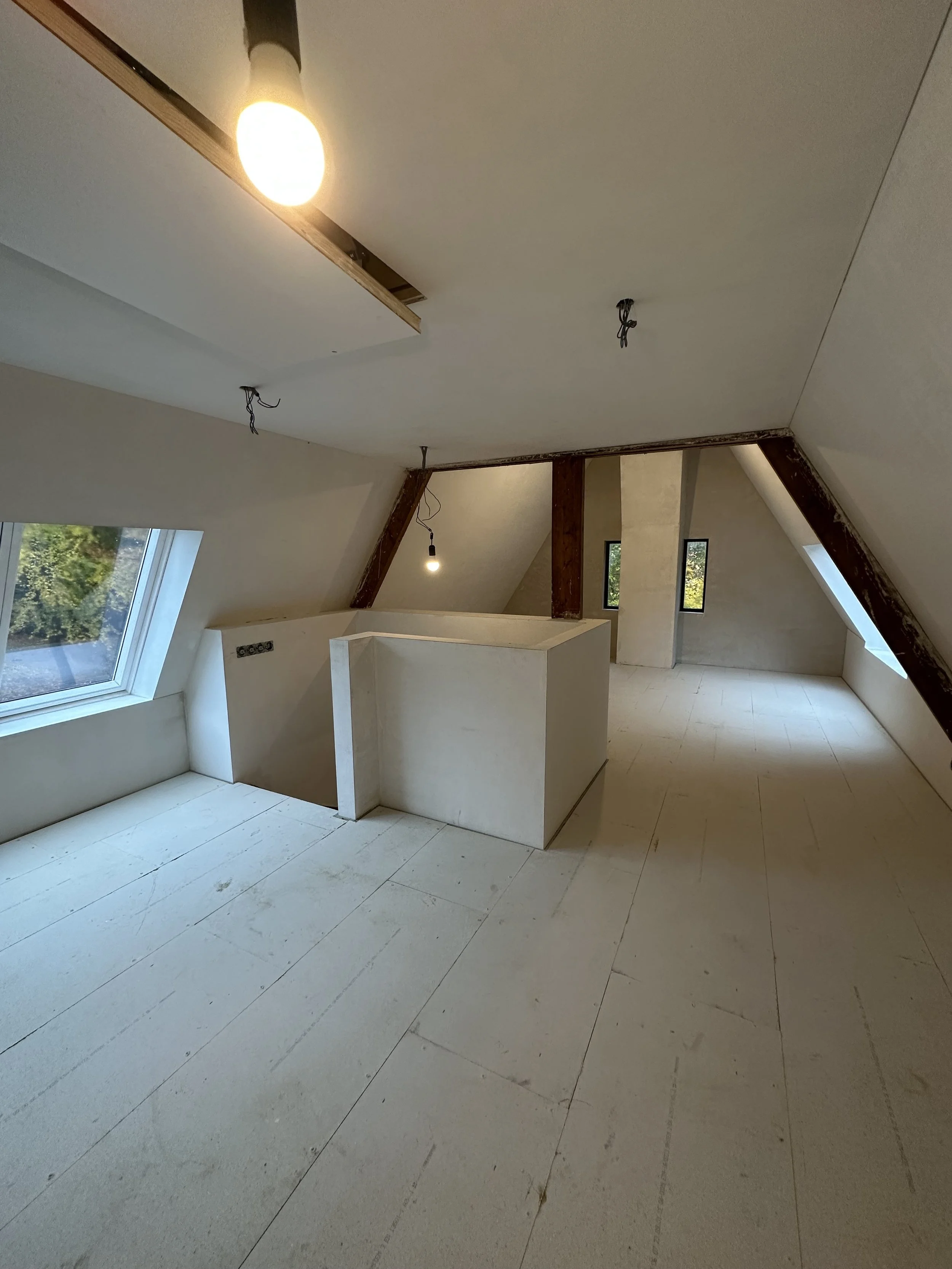 Attic or loft space under construction with unfinished flooring, sloped ceilings, exposed beams, and windows, with light fixtures hanging from the ceiling.