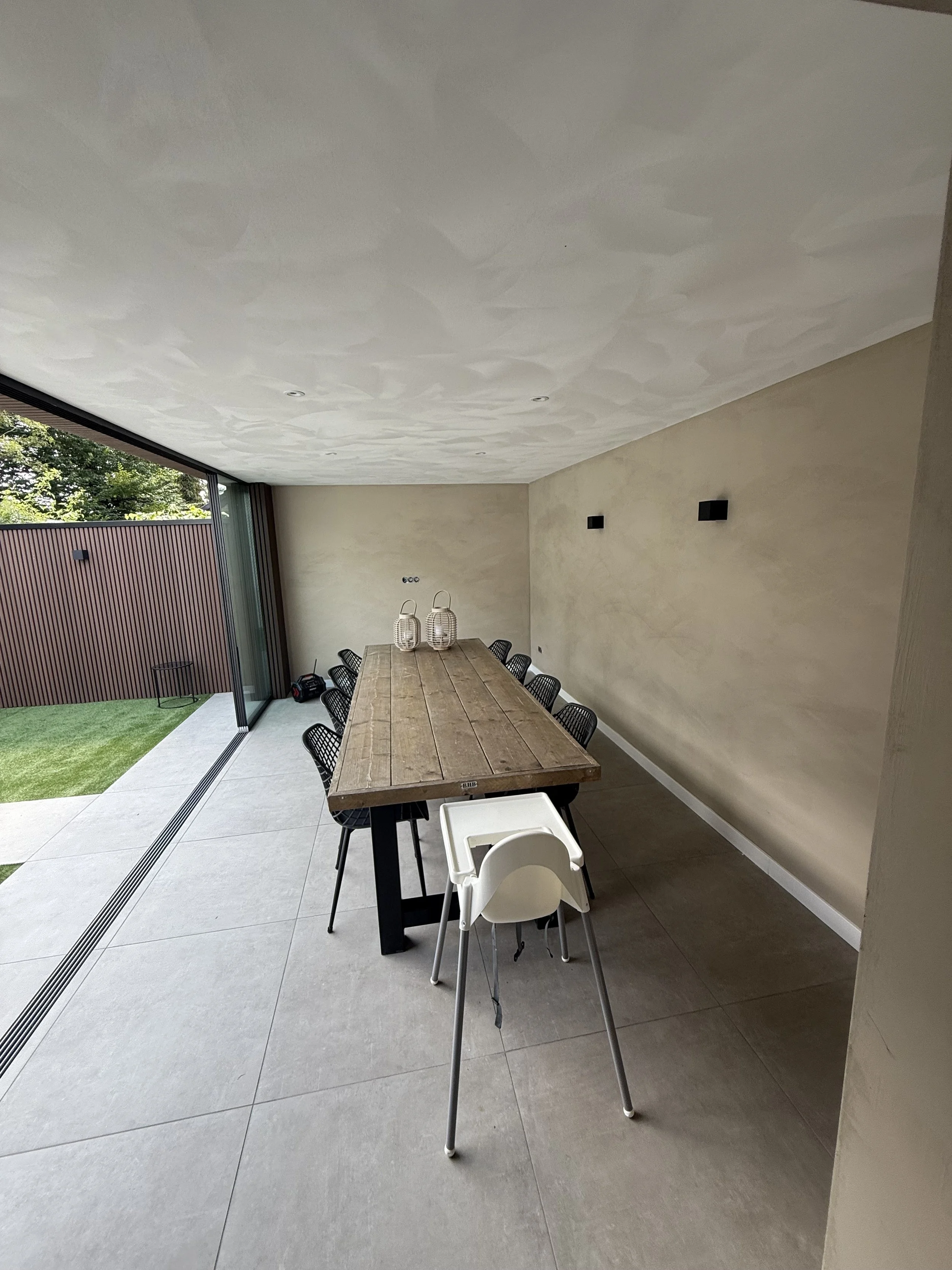 Modern dining room with large wooden table, black chairs, some decorative lanterns on the table, and a view of a backyard with grass and a wooden fence through sliding glass doors.