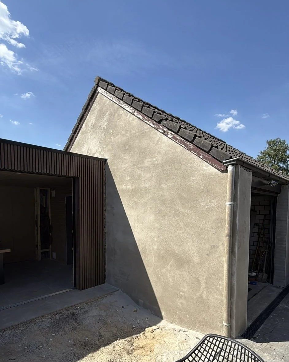 Side view of a building under construction with a smooth stucco wall, a corrugated metal exterior, a stone tile roof, and a rain gutter system, with a partly cloudy sky in the background.
