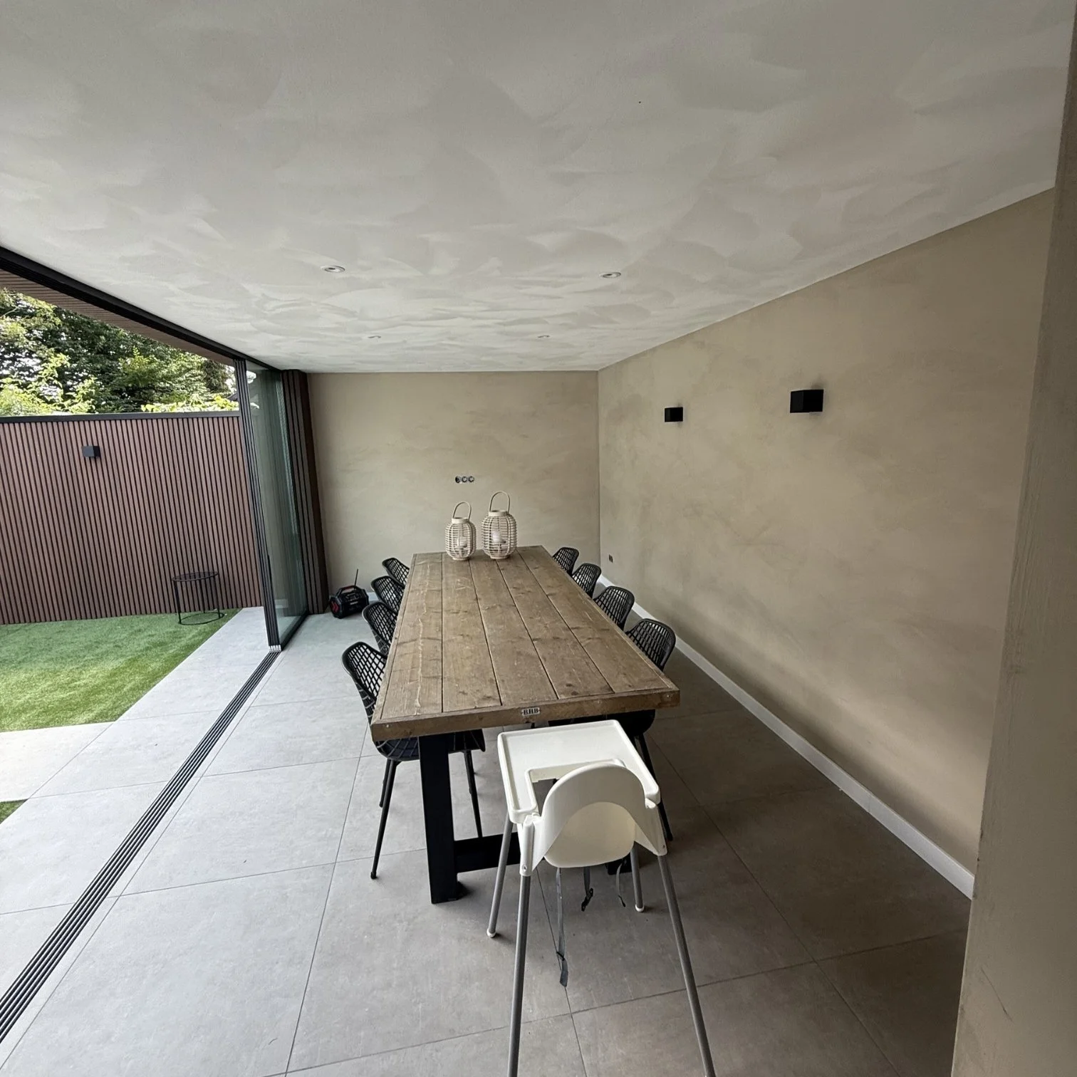 Modern dining area with a long wooden table, black chairs, and a child high chair, next to a large glass sliding door opening to a backyard with grass and a wooden fence.