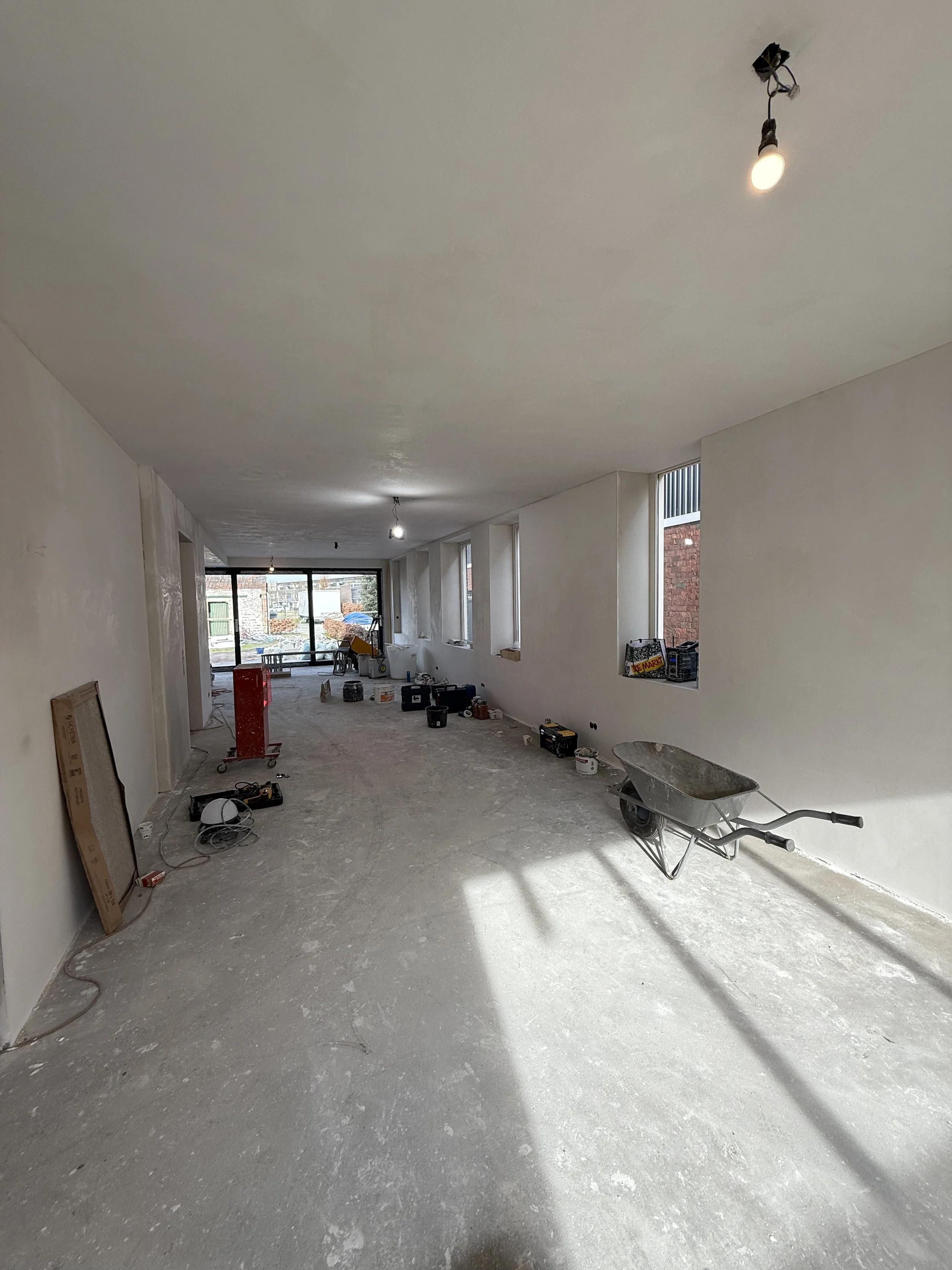 Interior of a room under construction with a wheelbarrow, construction tools, and supplies on the unfinished floor, and windows along one wall showing an outdoor view.