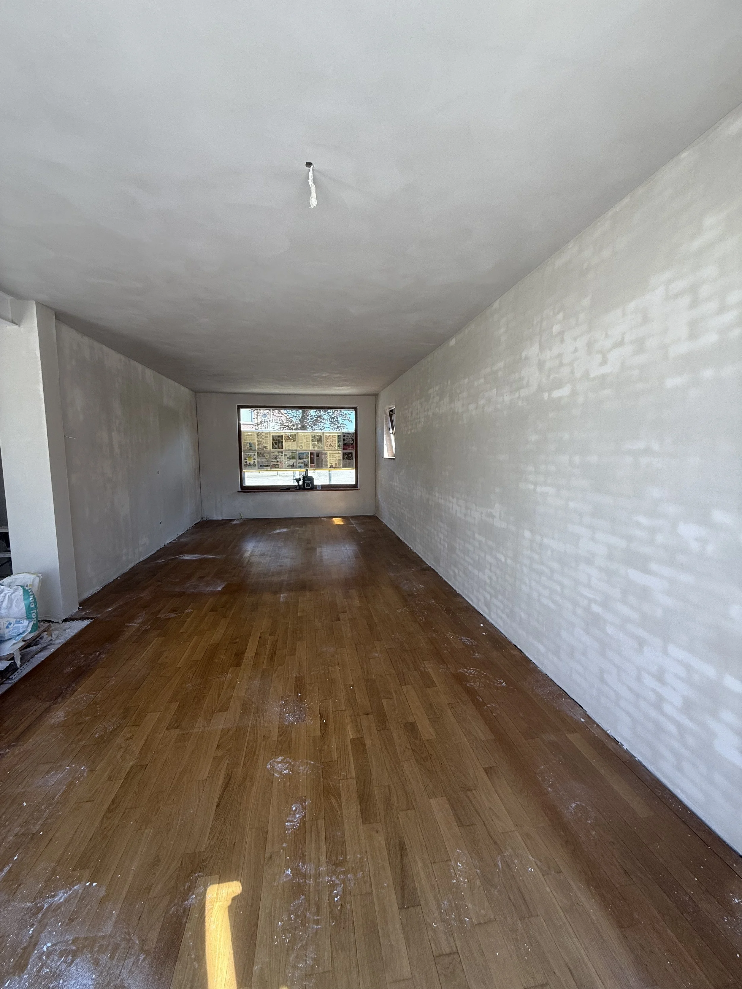 Empty room with white walls and wood flooring, under renovation, with a large window and a smaller side window.