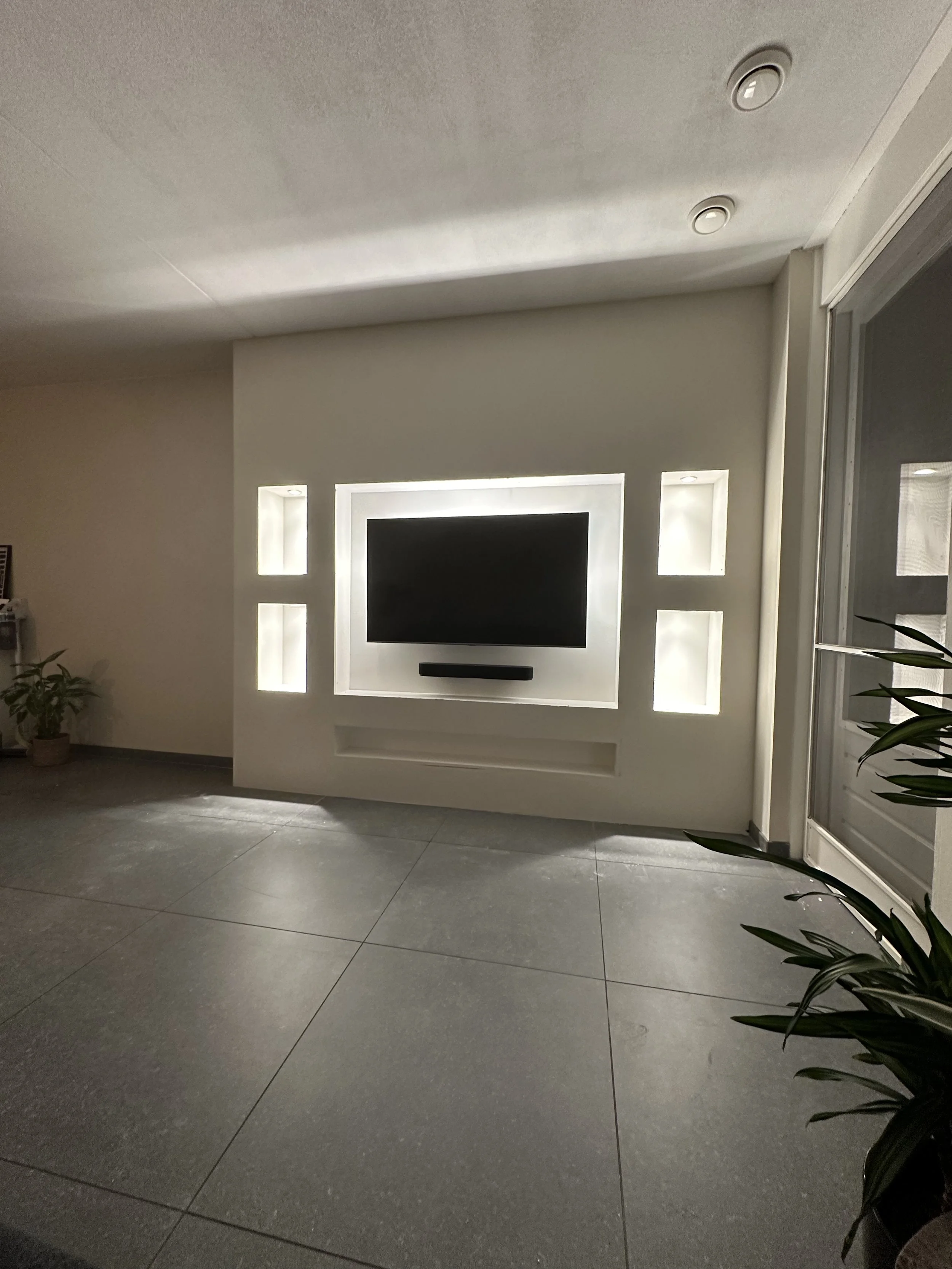 Living room with a wall-mounted flat-screen TV in a rectangular frame with backlit shelves on either side, and a soundbar below the TV. There are potted plants in the room, one on the left in a corner and another on the right near a sliding glass door.