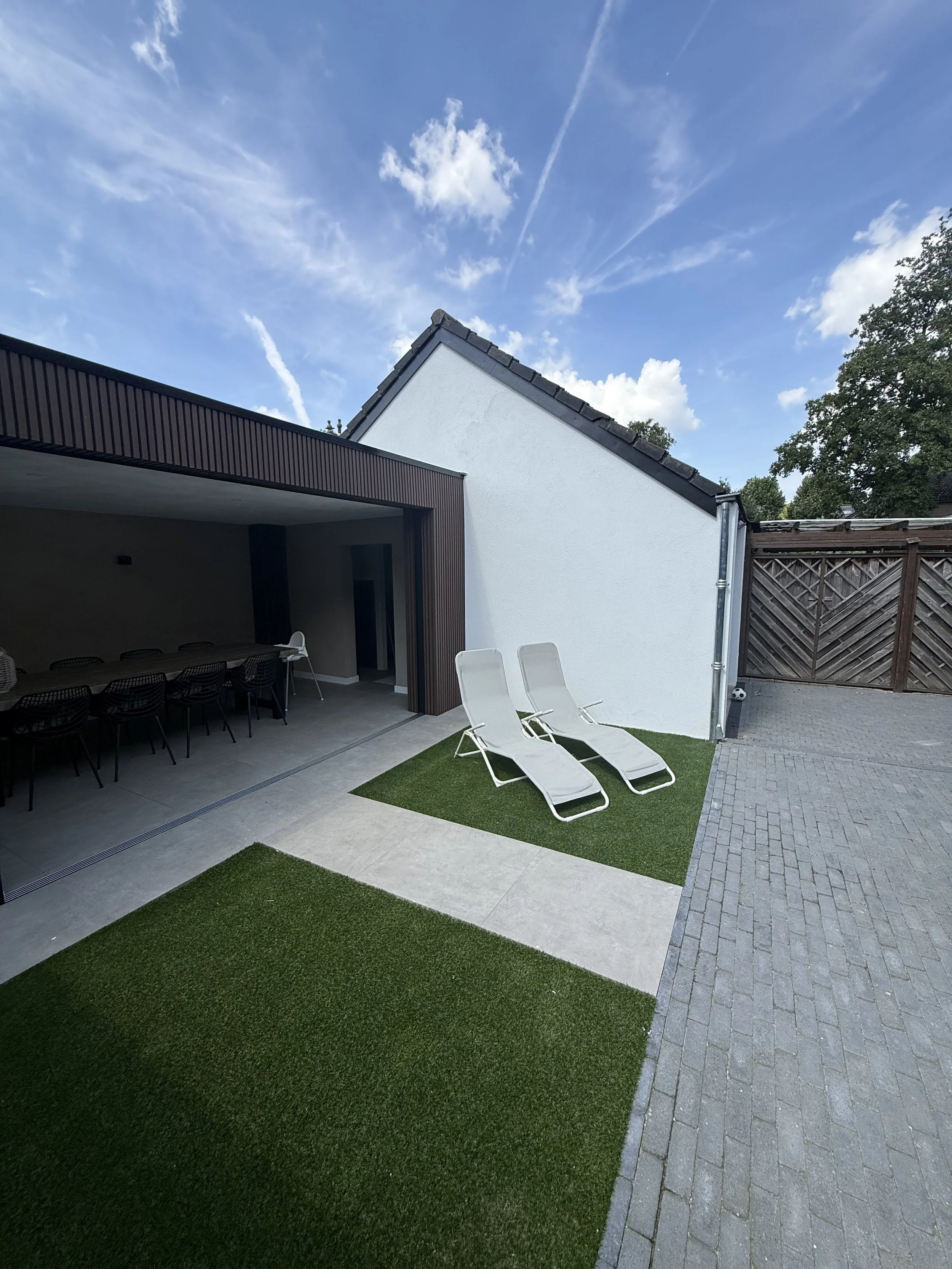 Modern backyard patio with two white lounge chairs on grass patch, adjacent to a gray brick path, next to a white house with dark roofing and a covered outdoor dining area.