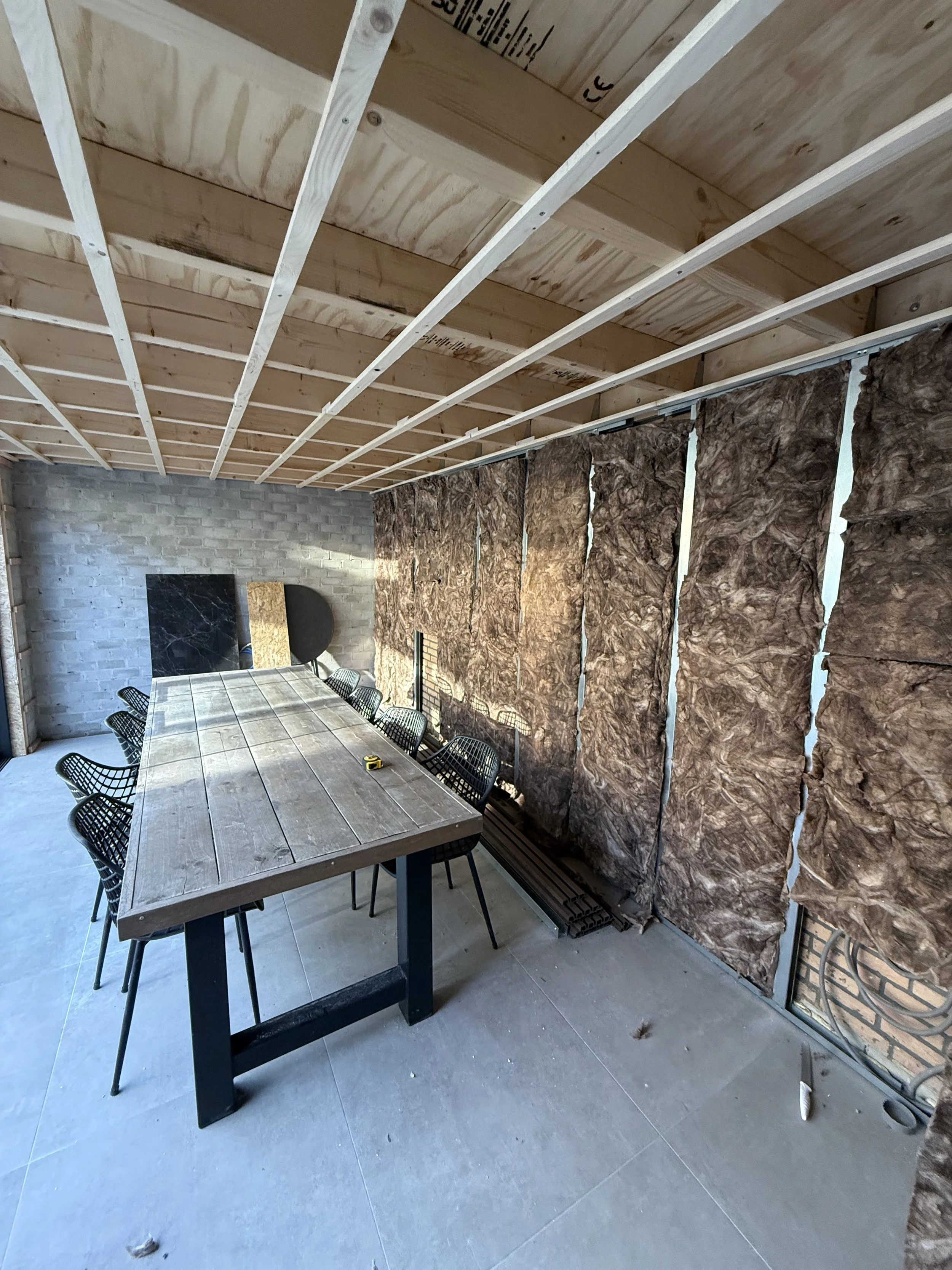 Construction of a room with insulation panels on one wall, a wooden ceiling frame, a large dining table, and black chairs.