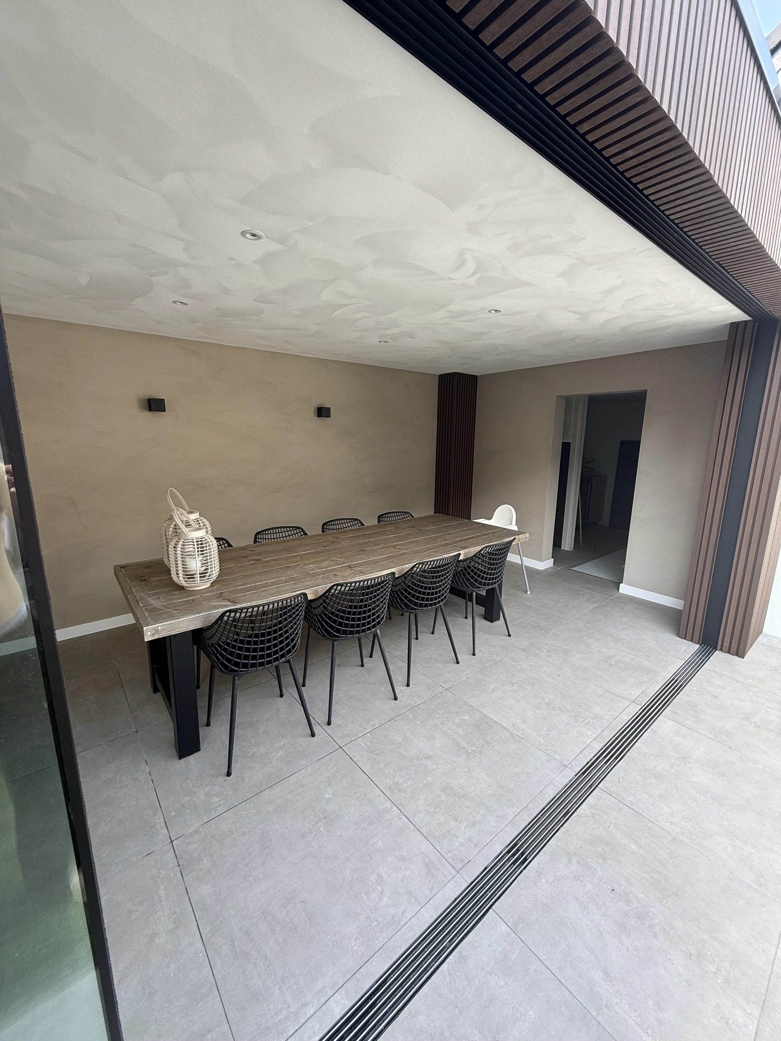 Modern outdoor patio with a large wooden dining table, eight black wire chairs, and a white high chair, under a ceiling with recessed lighting, surrounded by a beige wall and wooden accents.