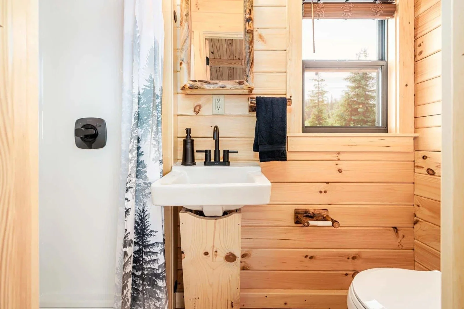 Small bathroom with wooden panel walls, a white sink with black fixtures, a mirror with a wood frame, a black towel, a window showing trees outside, and a shower curtain with a nature scene.