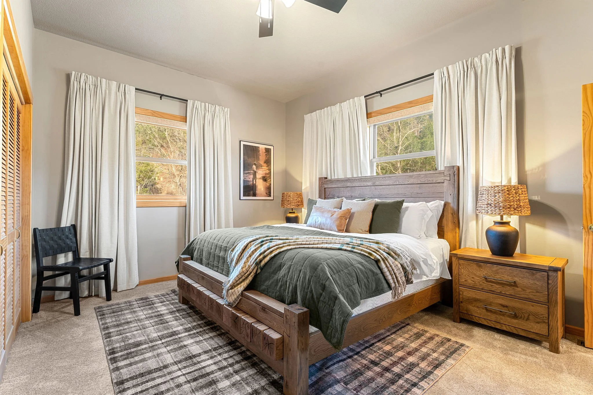 A cozy bedroom with a wooden bed frame, green and beige bedding, and multiple pillows, flanked by matching nightstands with lamps. The room features two large windows with white curtains and a painting on the wall, with natural light coming in.