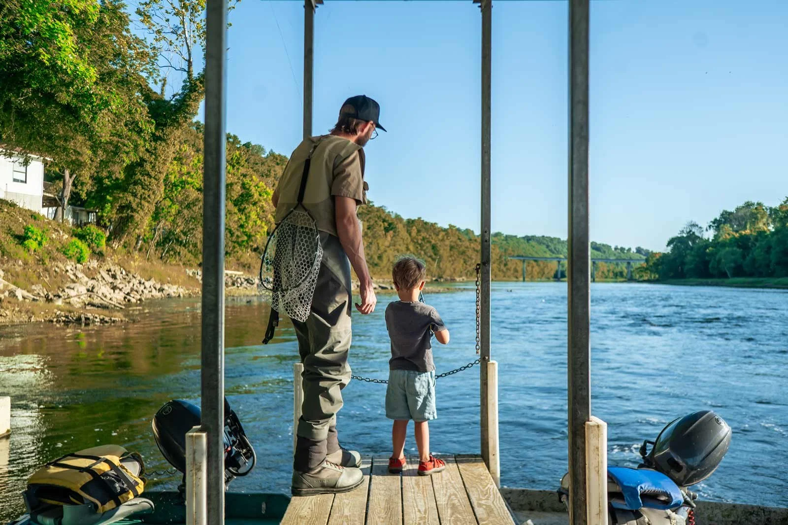 A man and a young boy stand on a dock by a river, fishing with a net. The scene is outdoors on a bright, sunny day with trees and a bridge in the background.
