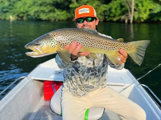 A man sitting in a boat holding a large fish with a green and black spotted pattern and a broad head. The man is wearing sunglasses, an orange cap, a camouflage long sleeve shirt, and beige pants. The background shows water and trees.