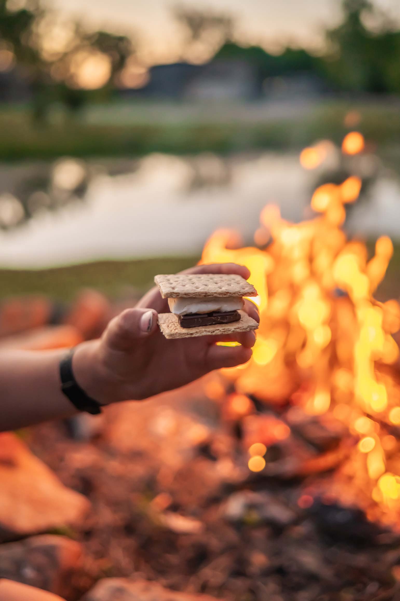 Person holding a s'more with a campfire and blurred outdoor background at sunset.