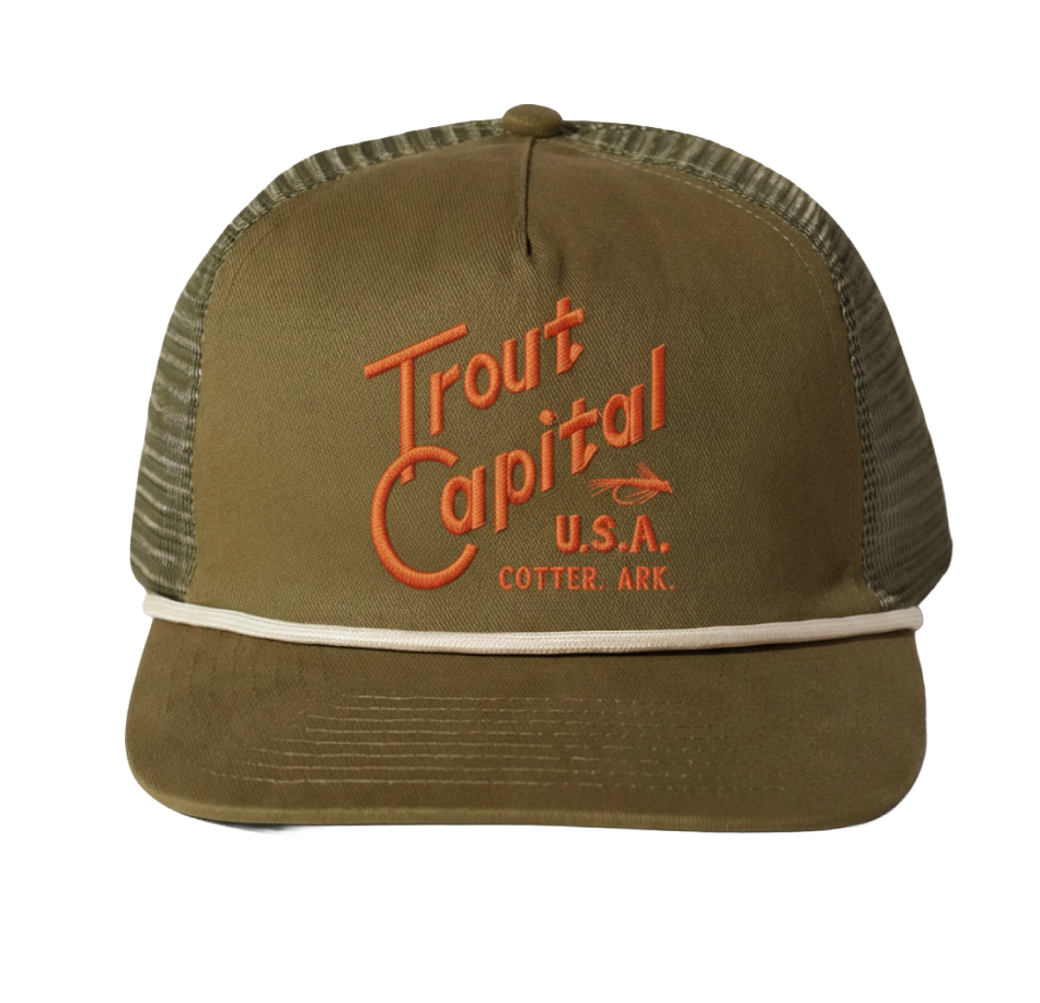 A khaki trucker hat with orange embroidered text reading 'Trout Capital U.S.A. Cotter, Ark.' and a small fish graphic.
