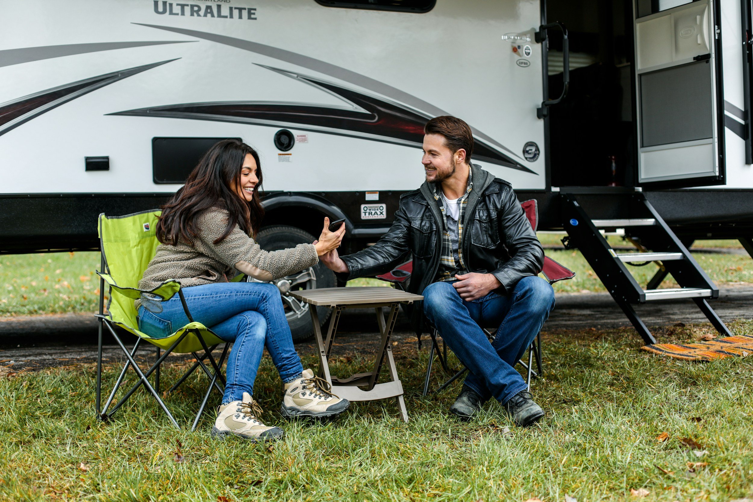 A man and woman sitting outside in front of an RV, smiling and looking at a phone together.