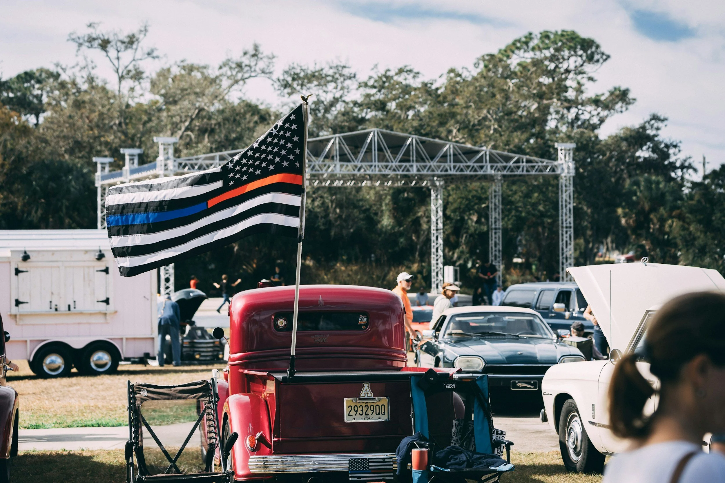 Black and white American flag with a thin red stripe, a thin blue stripe, and white stars on a flagpole at a car show, with tents, vintage cars, and a stage in the background.