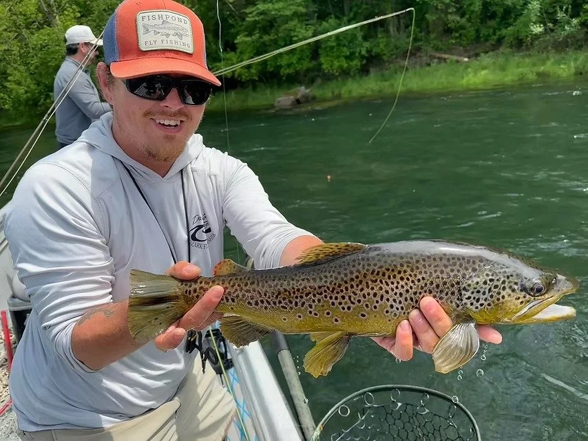 A man fishing from a boat holds a large brown trout with spots, smiling, while another person fishes in the background on a river surrounded by green trees.