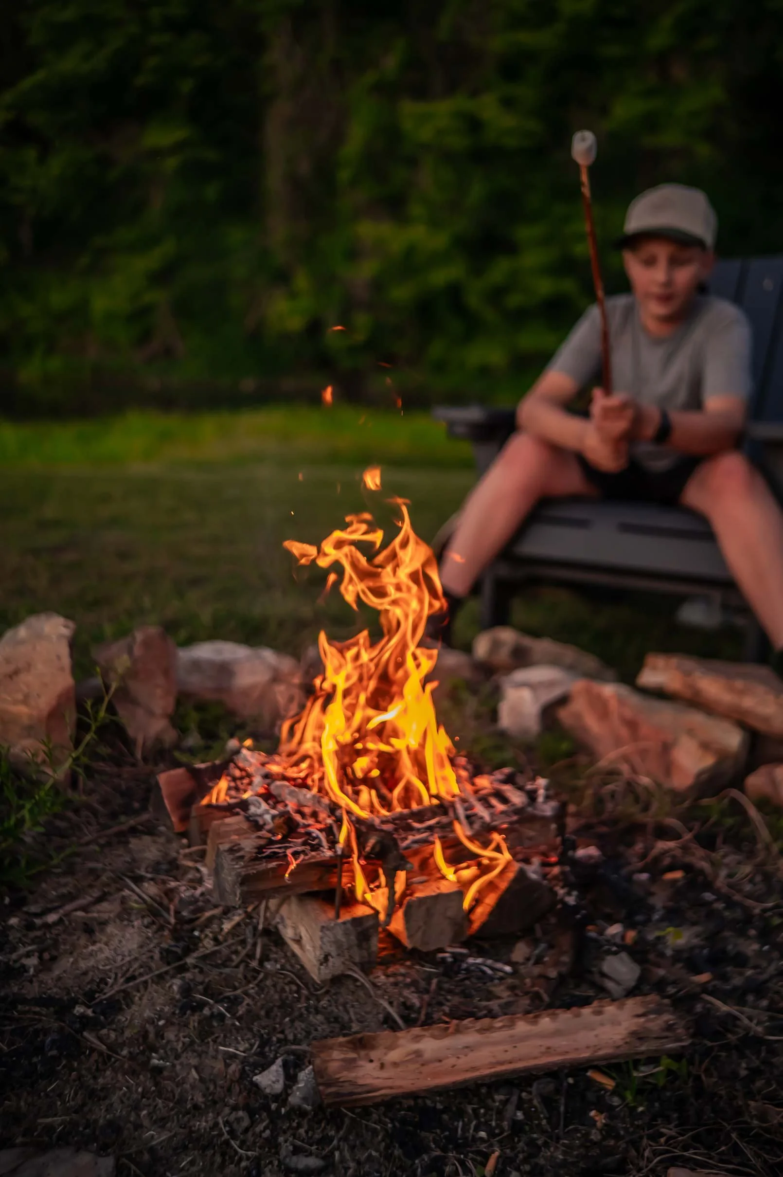 A boy sitting in a chair near a campfire, holding a stick with a marshmallow, in an outdoor setting with trees in the background.