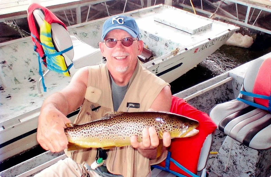 A man smiling and wearing sunglasses, a Kansas City Royals cap, and a vest, sitting in a boat and holding a large fish he caught.