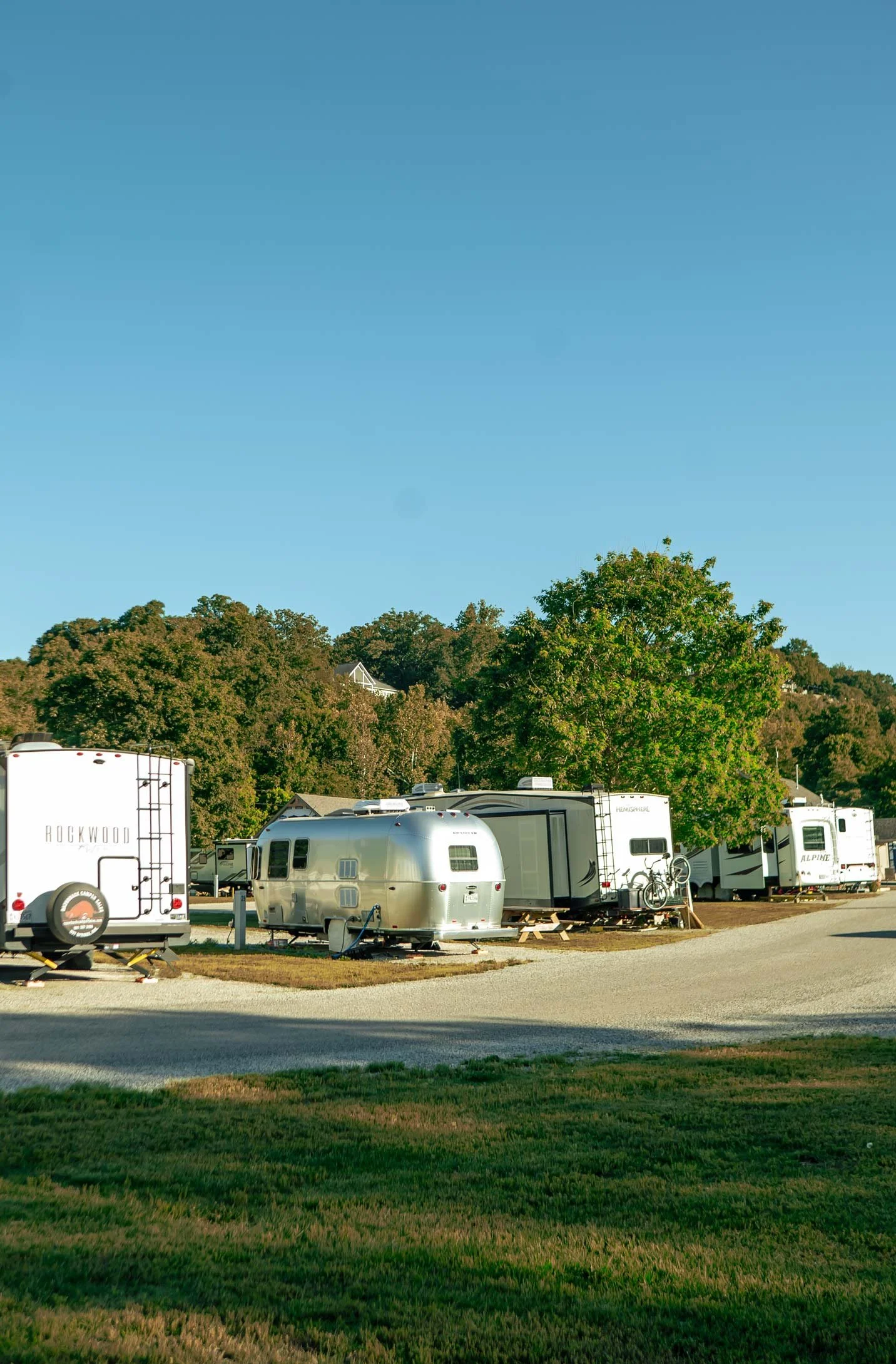 A parking lot filled with small travel trailers parked among green grass and trees, with a clear blue sky above.