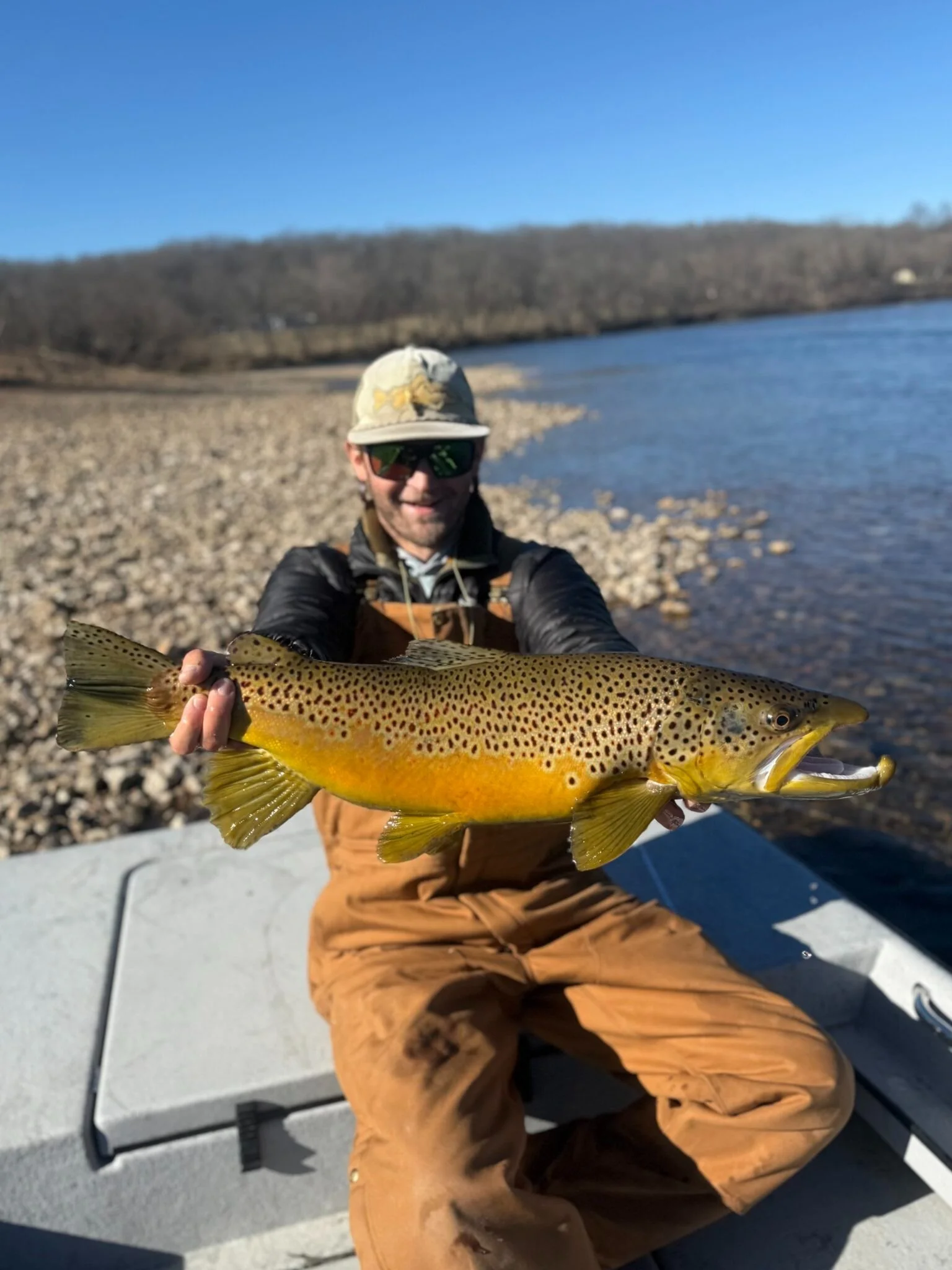 Man in outdoor clothing, wearing sunglasses and a hat, sitting on a boat and holding a large fish with a yellow and brown spotted pattern, by a river on a sunny day.