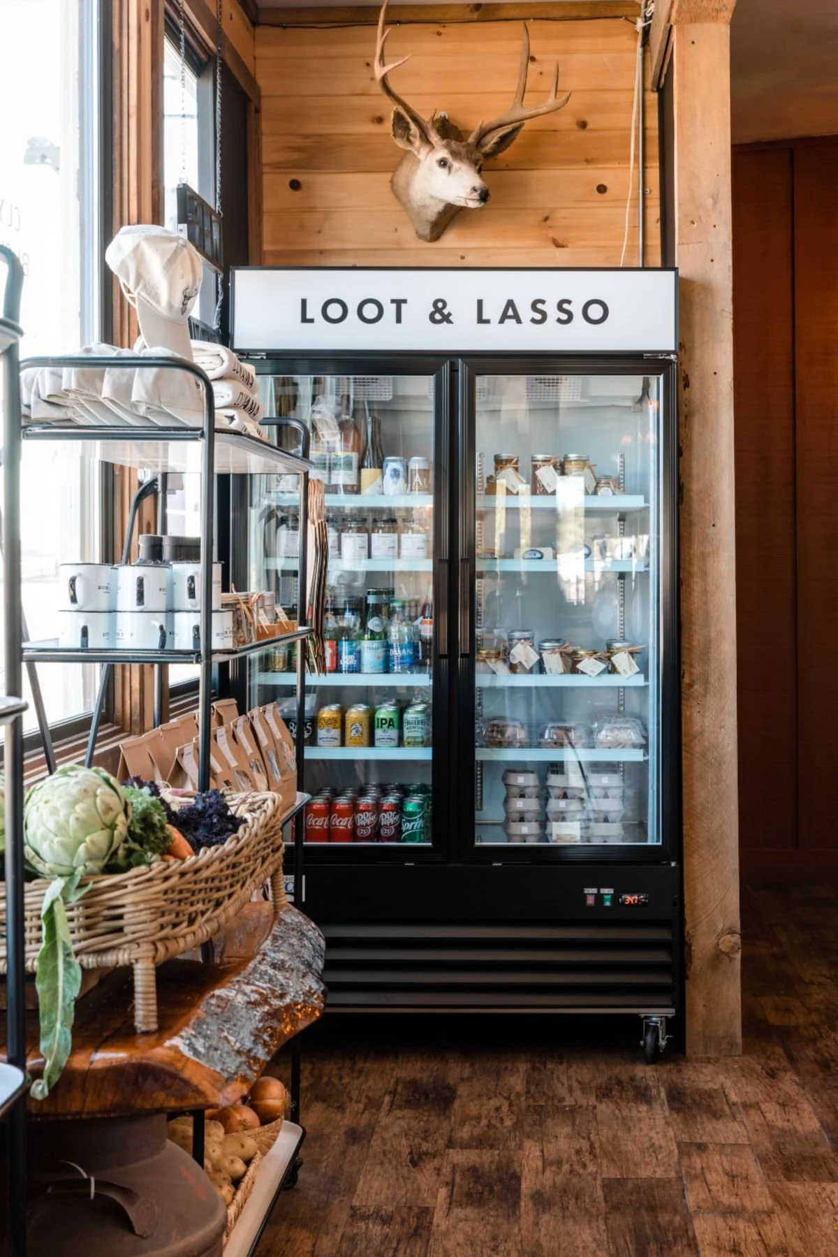 A wooden interior with a mounted deer head above a refrigerator, which has a sign reading 'Loot & Lasso'. The refrigerator contains beverages and food items, and there is a metal shelf on the left with clothing and other items, alongside a basket of vegetables on a rustic wooden table.