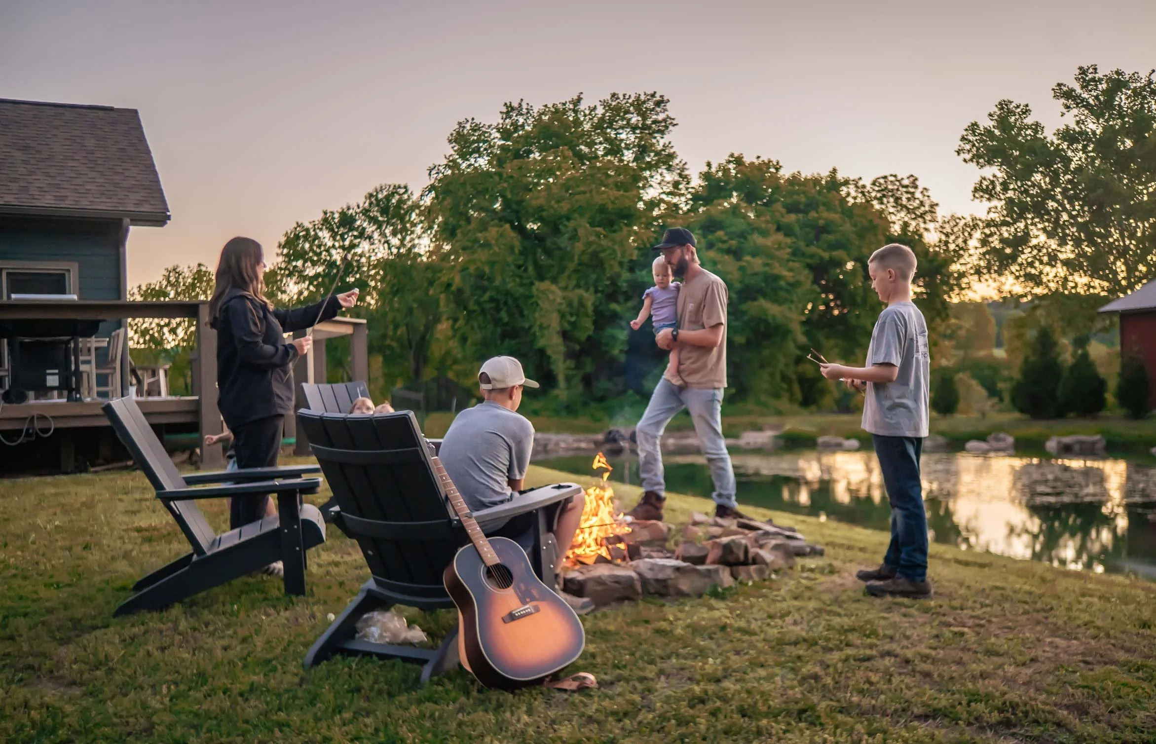People gathered around a small fire by a lake during sunset, with trees and a house in the background, some holding guitars and others watching.