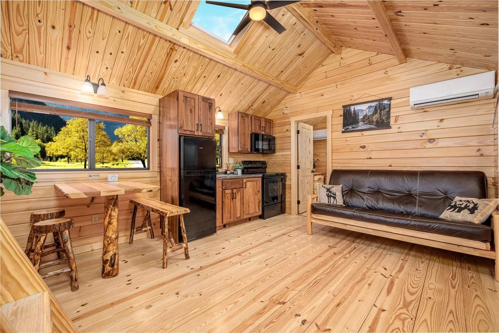 Interior of a cozy wood cabin with a vaulted ceiling, featuring a small living area with a brown leather sofa, a wood-paneled wall with a landscape picture, a black refrigerator, a stove, microwave, wooden cabinetry, a window showing a scenic outdoor