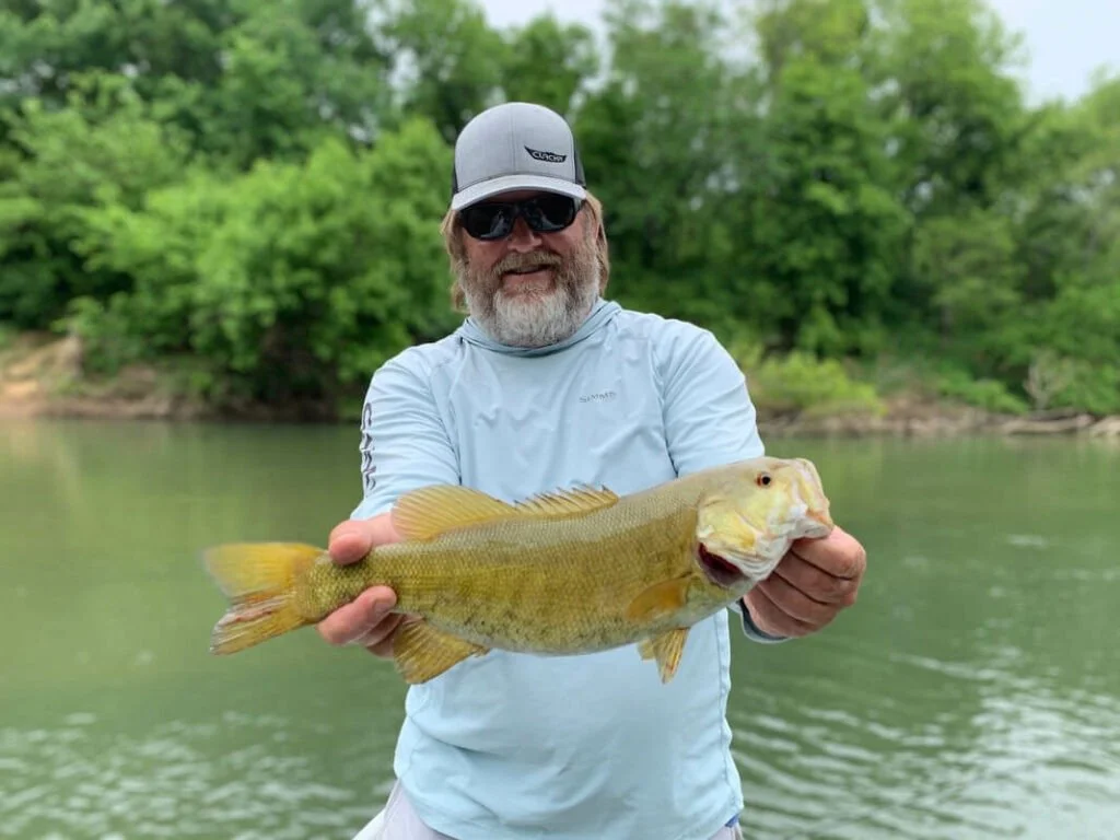 Man with gray beard, wearing sunglasses, a gray baseball cap, and light blue shirt, holding a large fish with a greenish-yellow body, near a river with green trees in the background.