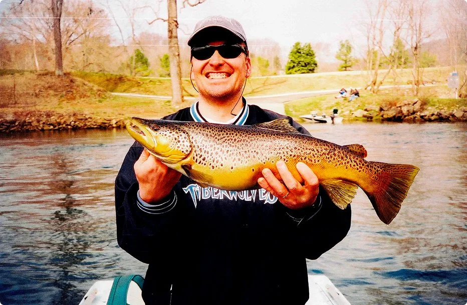 Man outdoors holding a large trout fish, smiling, near a river with trees in the background.